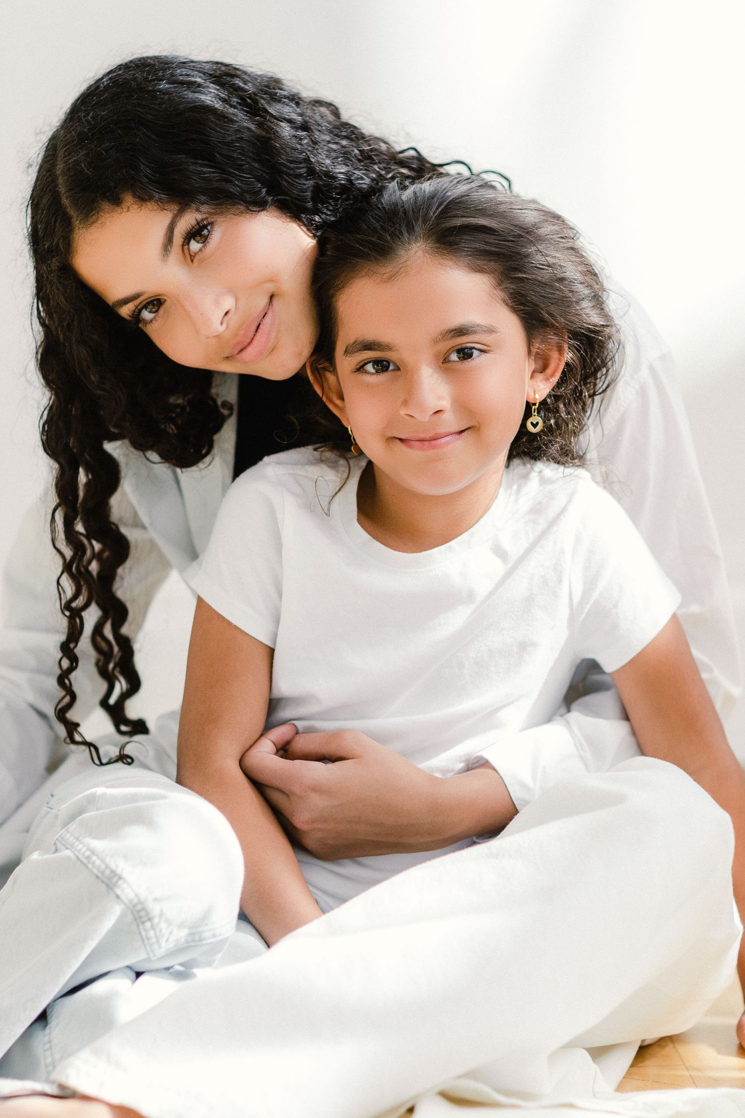 A woman with curly dark hair embraces a young girl in white outfit, both smiling warmly in a bright setting.