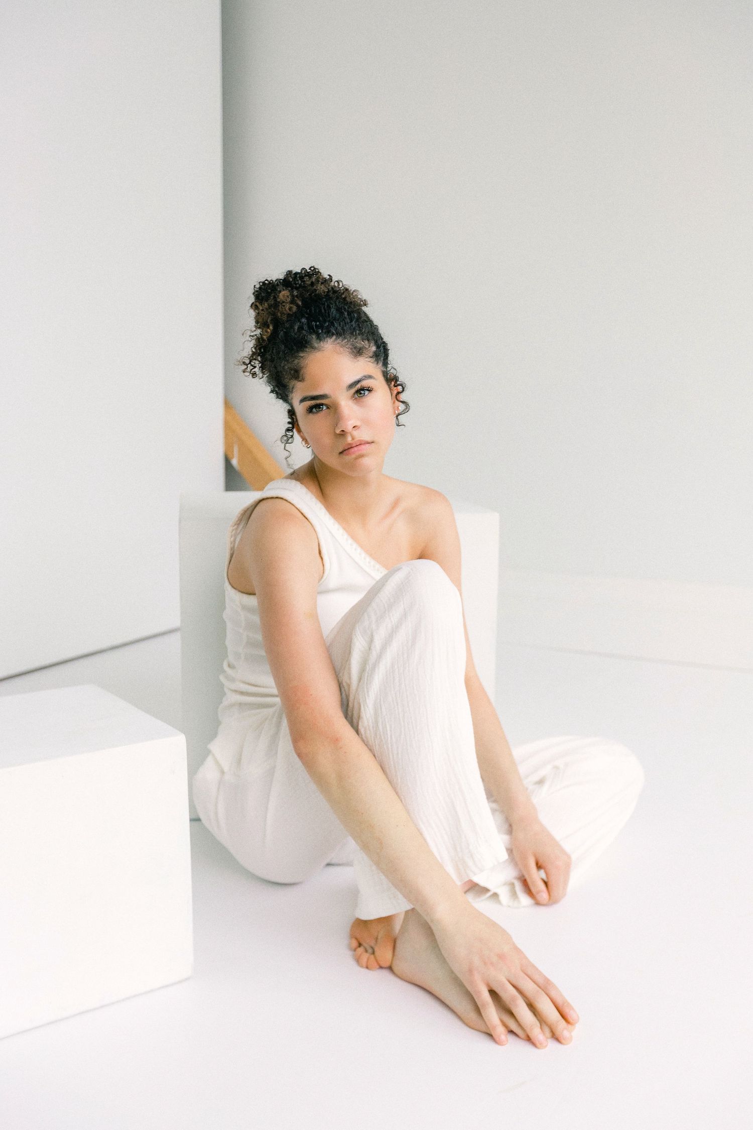Young woman in white one-shoulder top and pants sitting barefoot on white floor in minimalist studio.