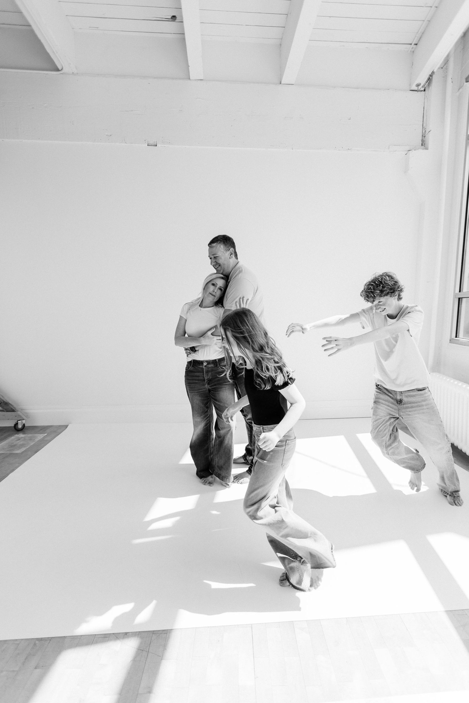 A lively black and white family photo with four people playing and embracing in a bright studio.