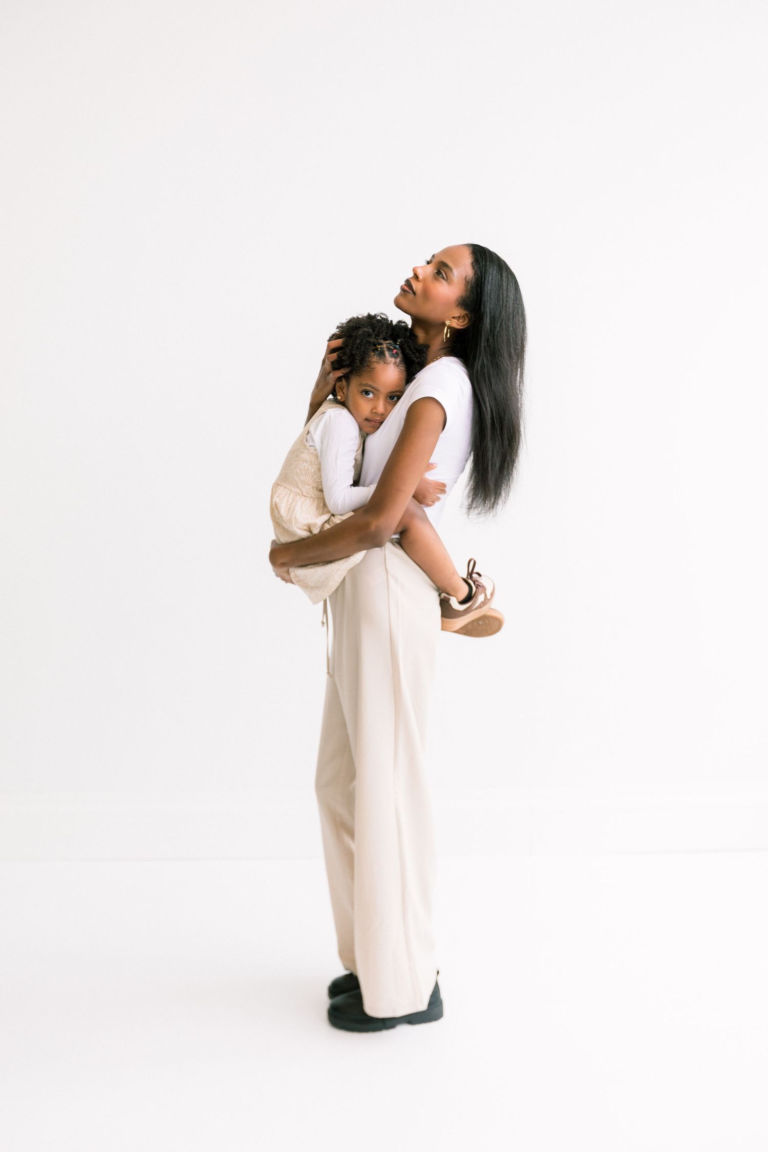 A mother her young daughter close while standing against a white background.