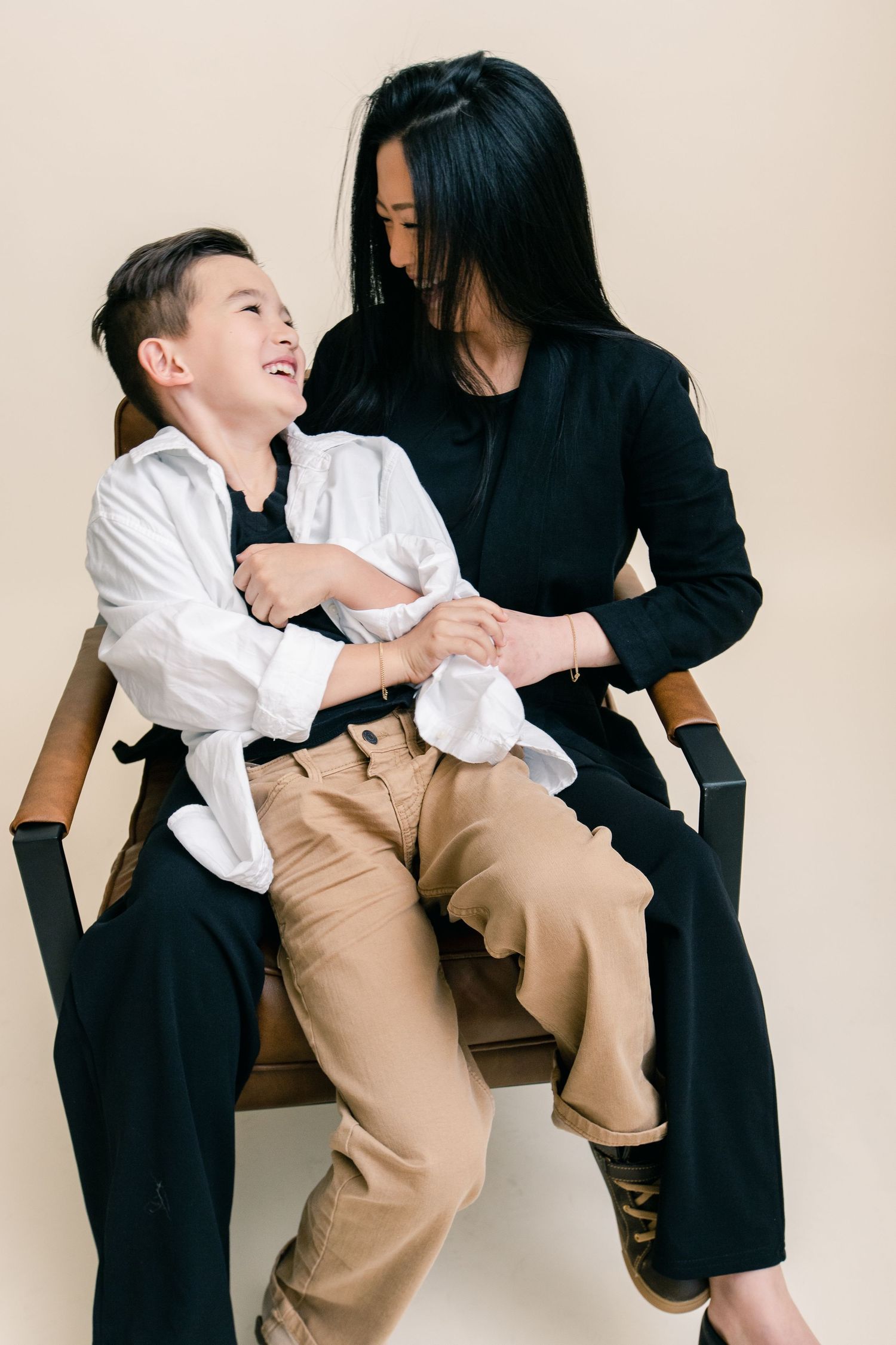 A woman in black sits on a chair with a laughing boy in a white shirt and tan pants on her lap.