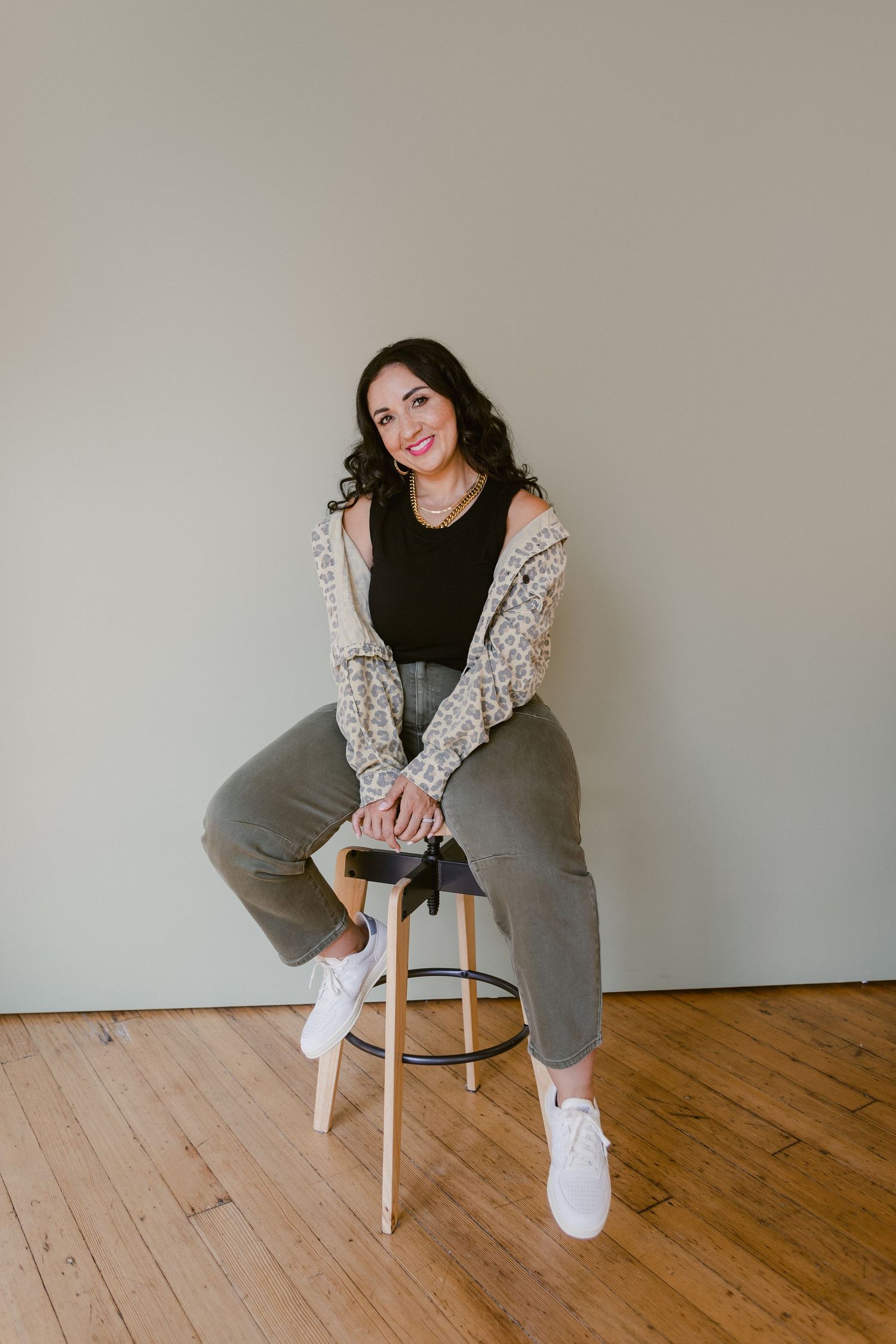 Woman in leopard cardigan and gray pants sitting on a wooden stool, smiling in a bright studio.