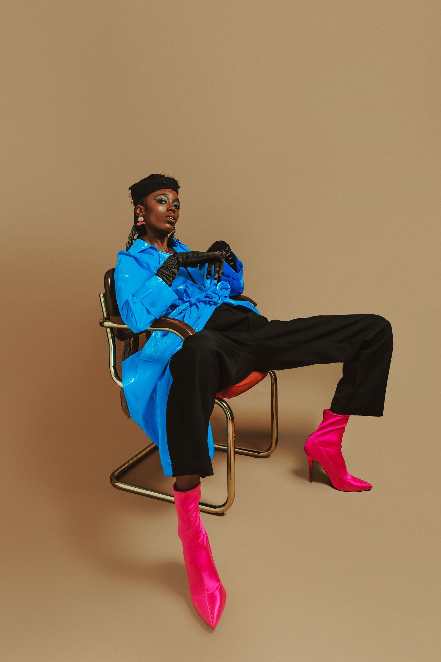 Stylish woman in blue jacket, black pants, and pink boots sitting on gold chair against beige background.
