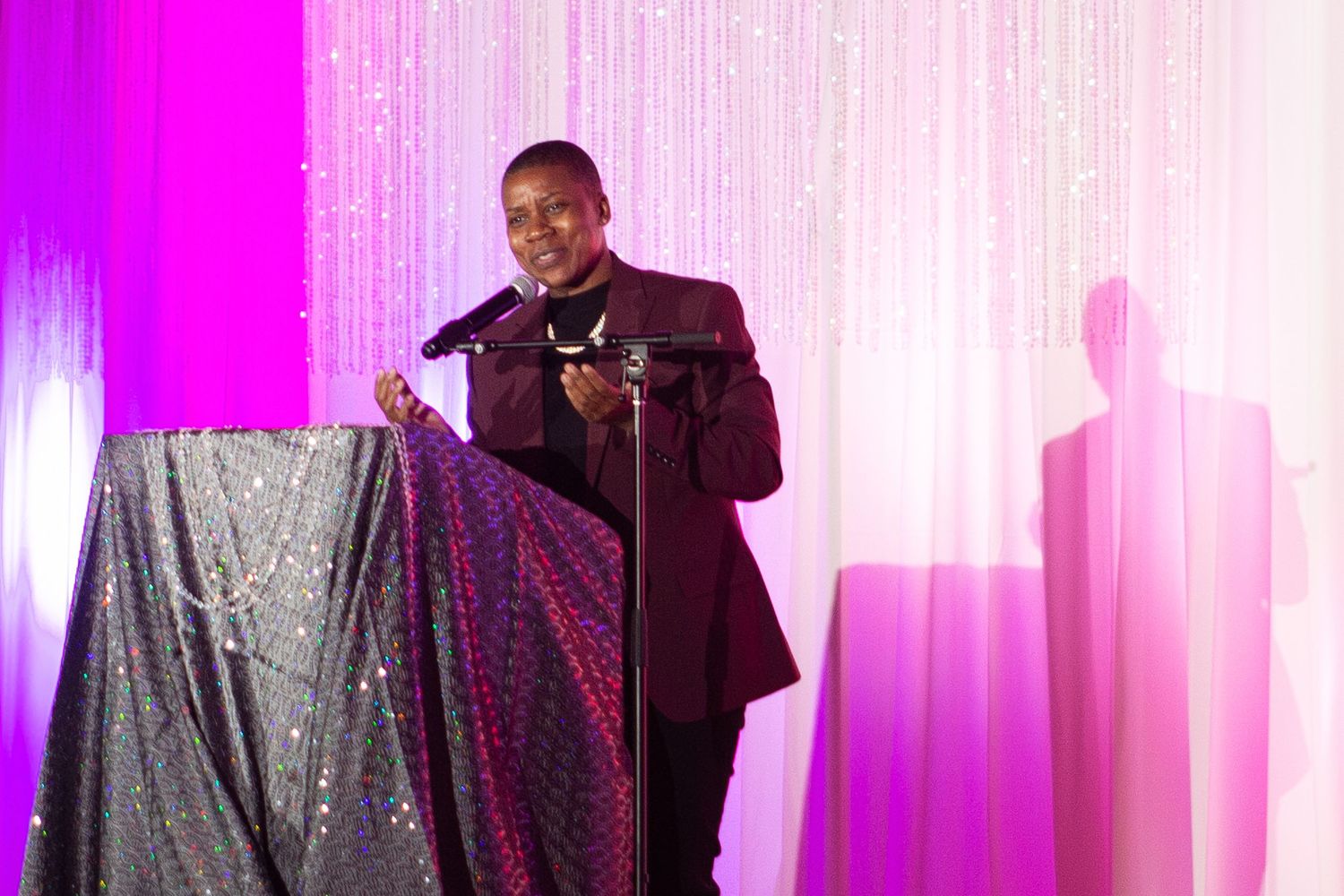 A man in a burgundy suit speaks at a microphone on stage with a sparkling backdrop and purple lighting.