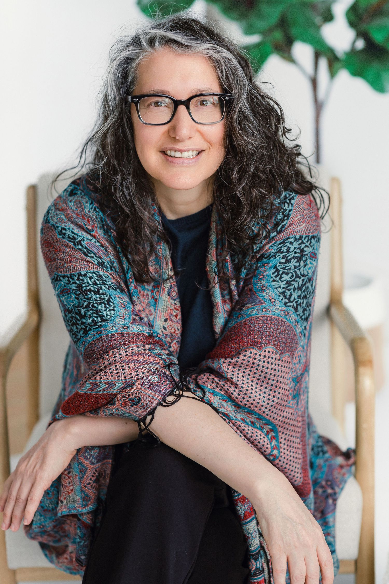 Smiling middle-aged woman with curly gray hair, glasses, wearing colorful patterned shawl, sitting relaxed in wooden chair.