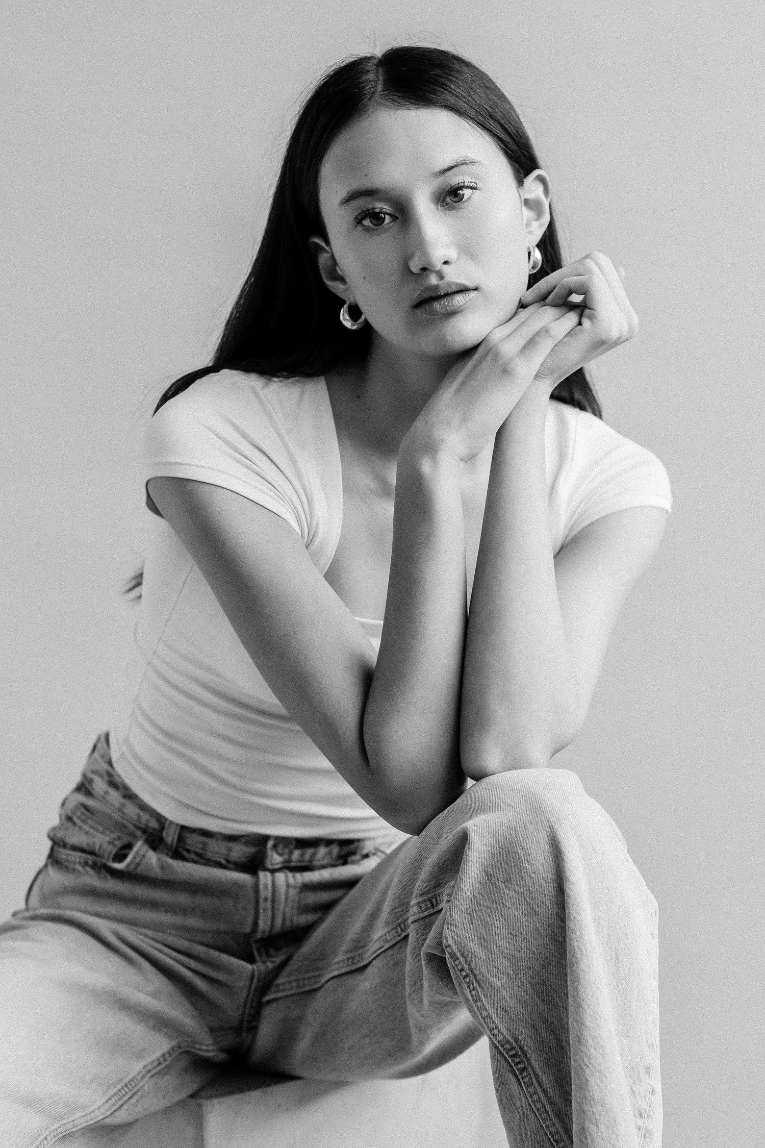Black and white portrait of a young woman in white t-shirt and jeans, posing thoughtfully with hands near face.