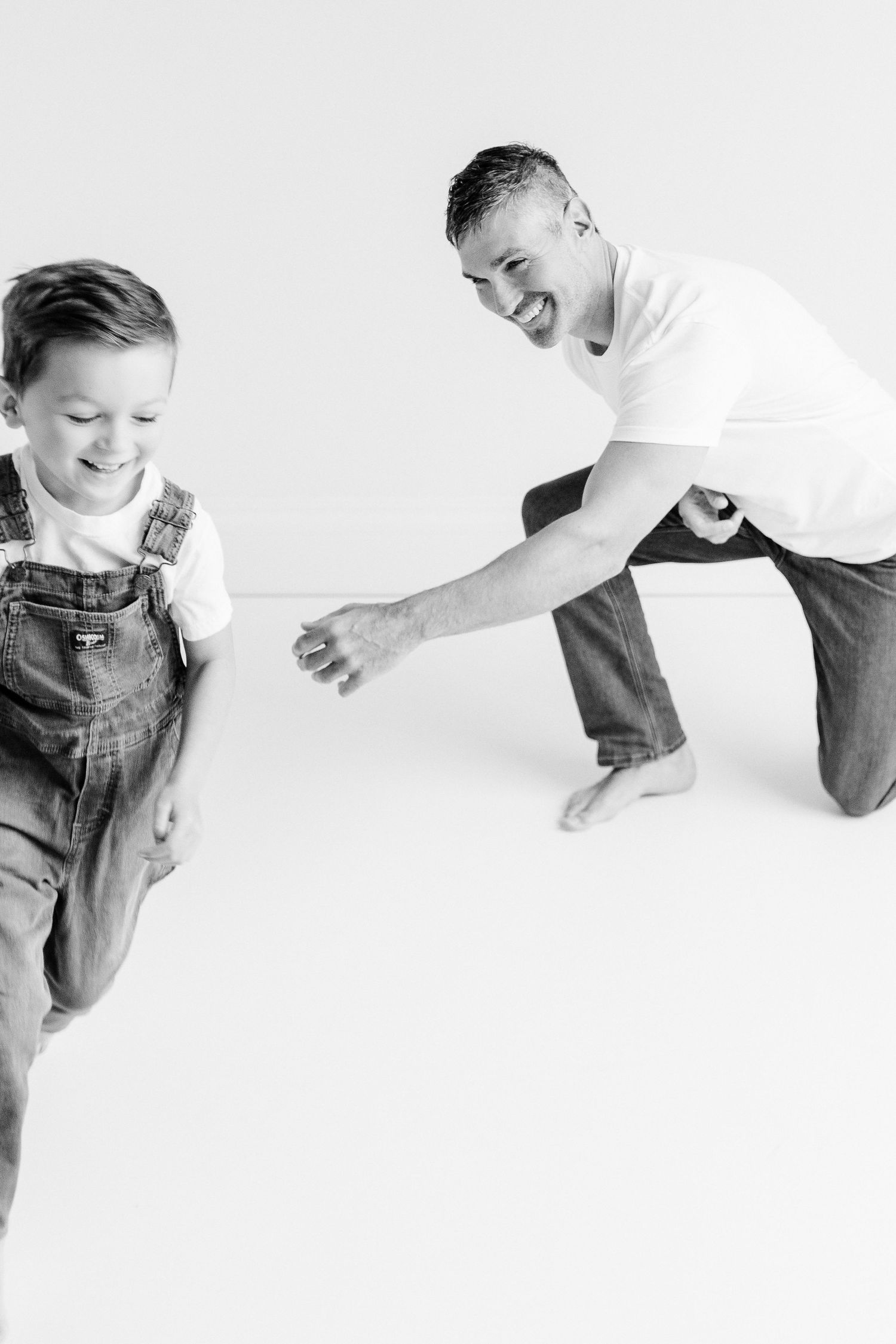 Black and white photo of a laughing father chasing his smiling young son wearing overalls in a bright studio.