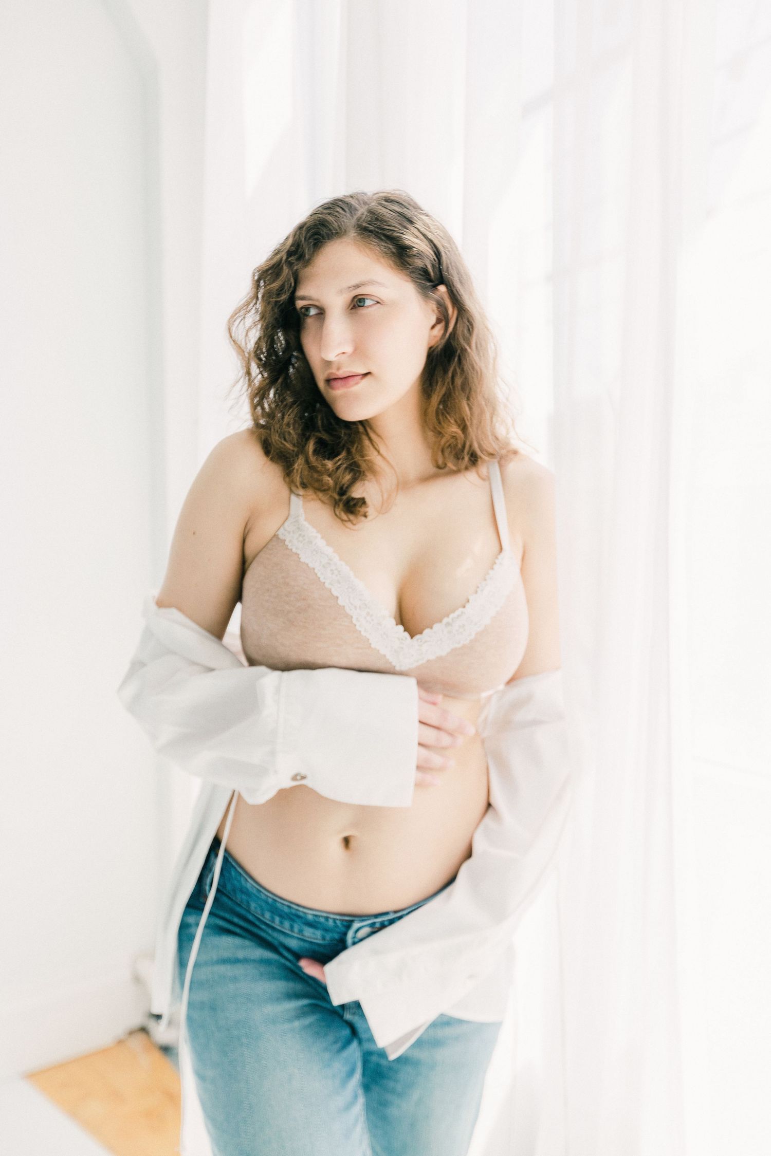 Young woman in beige lace bralette, jeans, and open white shirt posing near bright window in natural light.