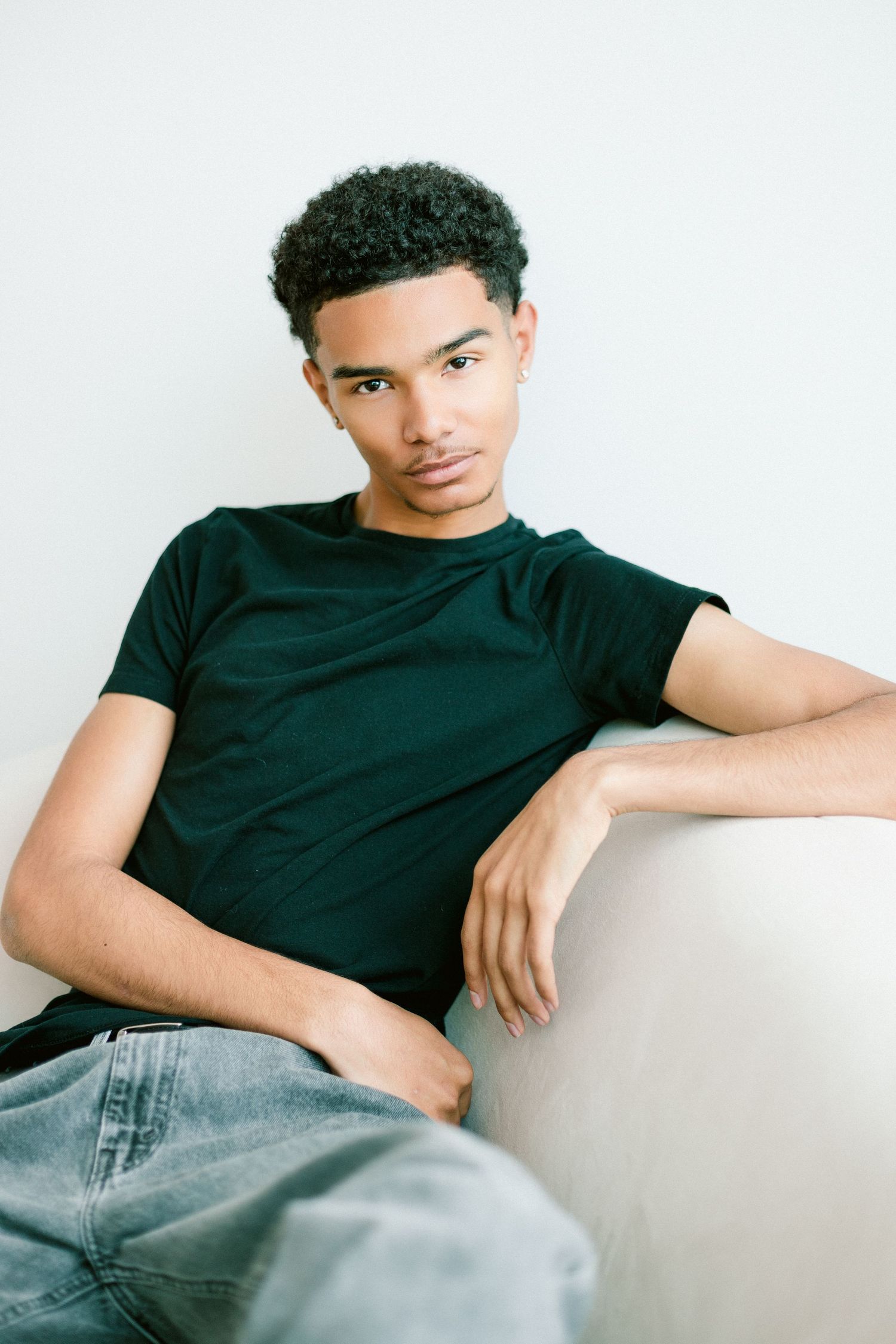 Young man in black t-shirt and gray jeans sitting relaxed on a white couch against a light background.