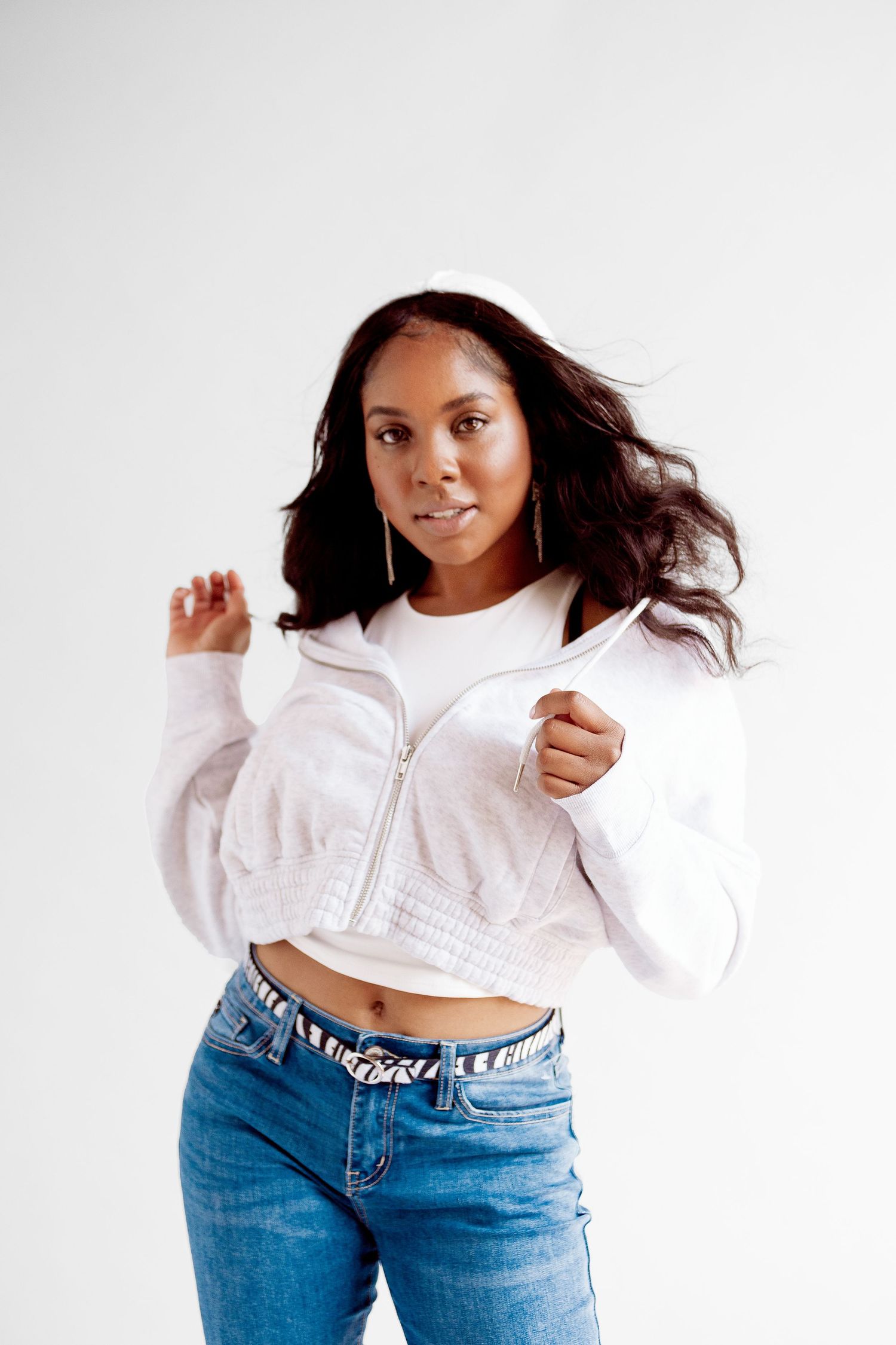 Young woman in white cropped zip-up jacket and blue jeans posing confidently against a white background.