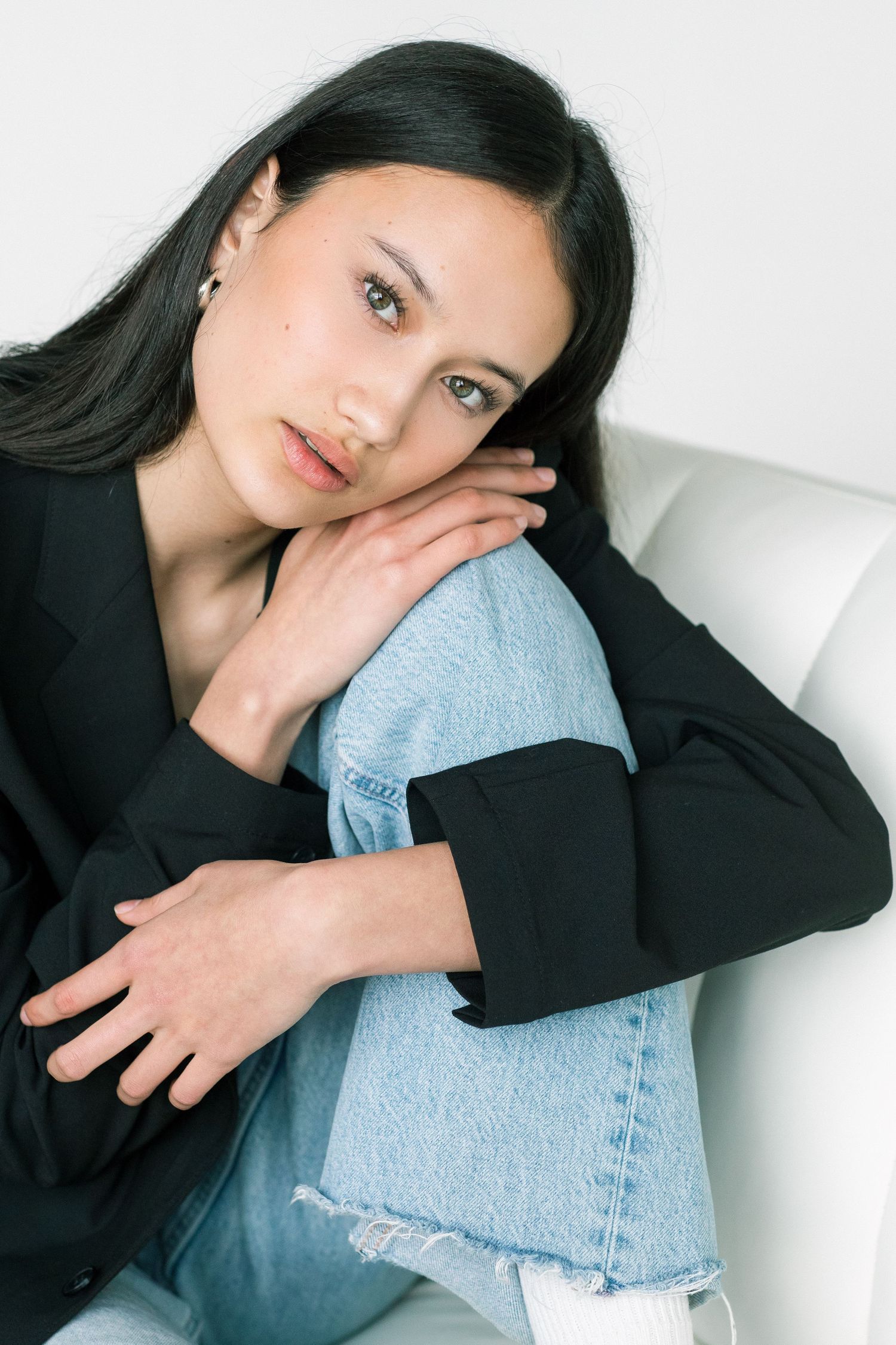 Young woman in black blazer and ripped jeans sitting relaxed on white chair, resting chin on knee.