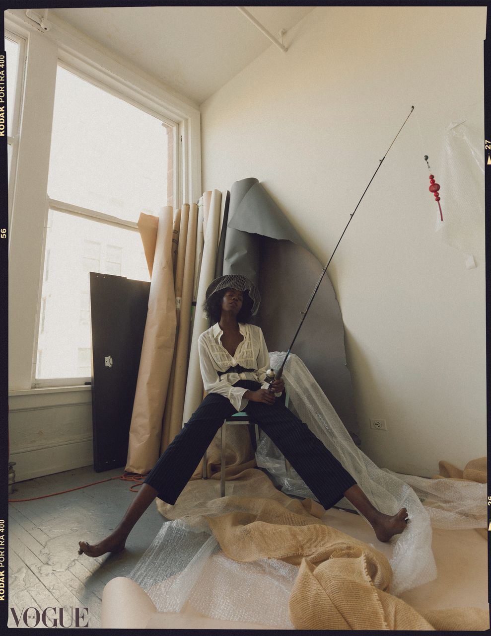 Vogue editorial portrait of a model seated in a studio space holding a fishing rod among draped fabrics.