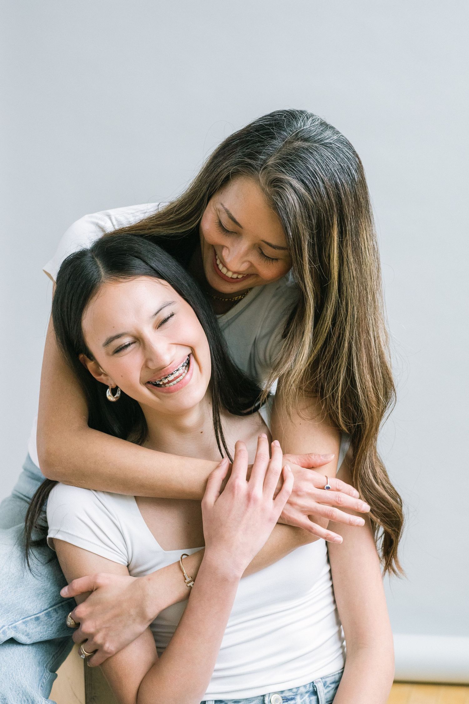 Two young women share a joyful hug, laughing together against a light gray background.