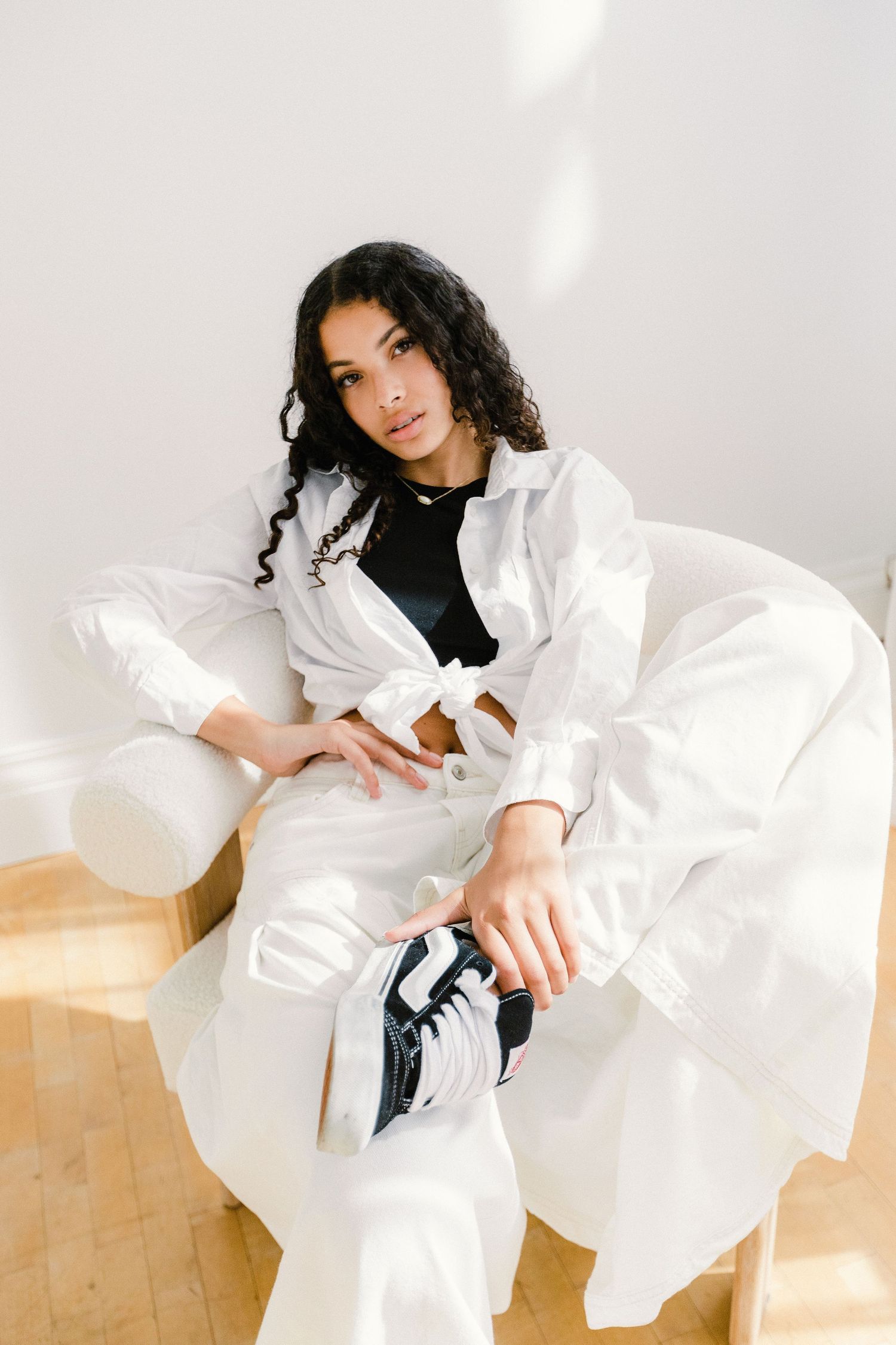 Woman with dark curly hair in white shirt and pants sits in white chair with sneakers.