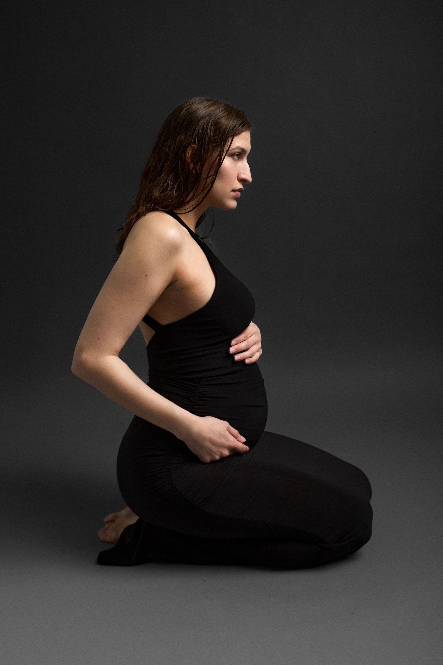 Pregnant woman in black outfit kneeling on floor, cradling baby bump, side profile view against dark gray background.