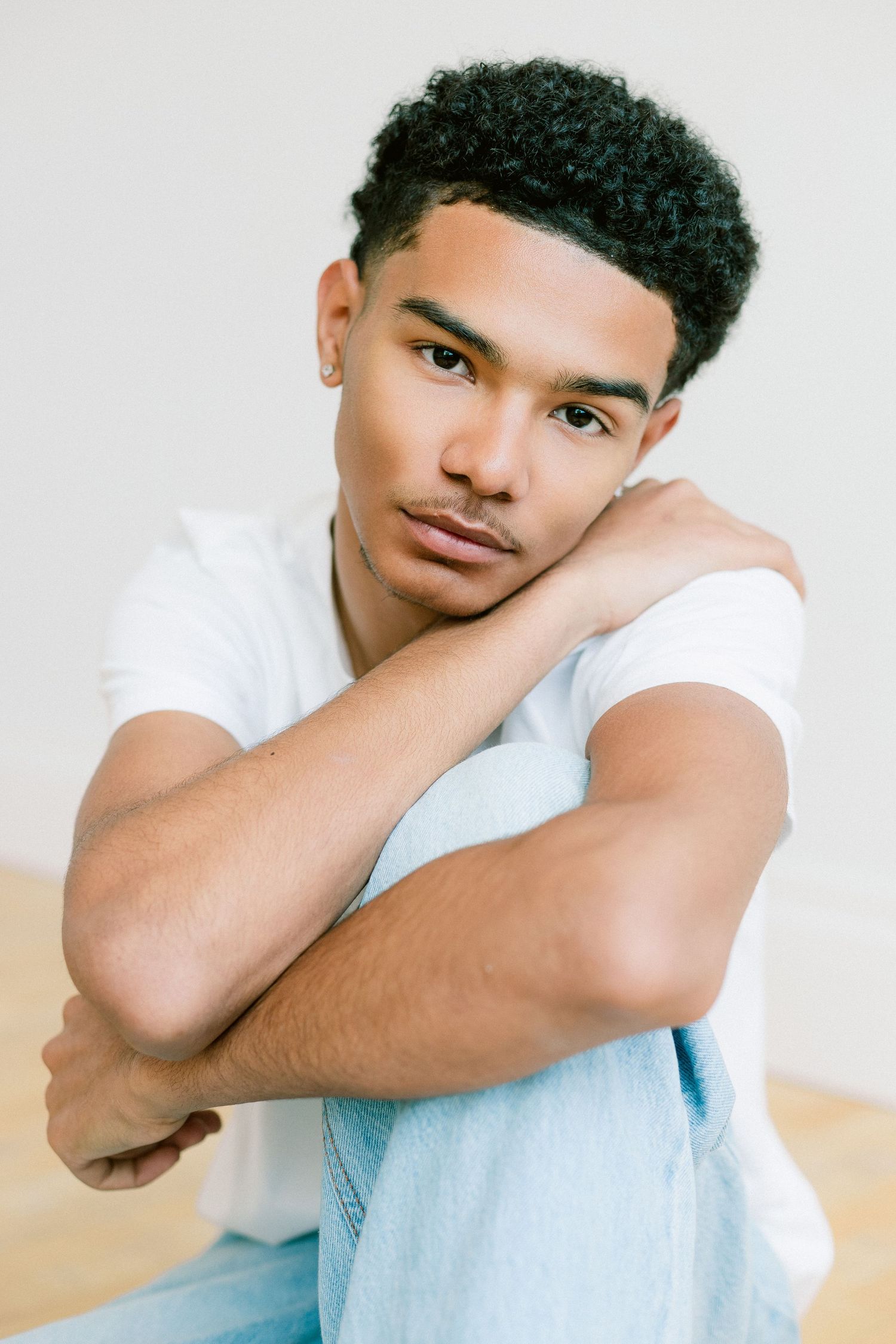 Young Black man in white t-shirt and jeans sits relaxed, resting chin on crossed arms, gazing at camera.