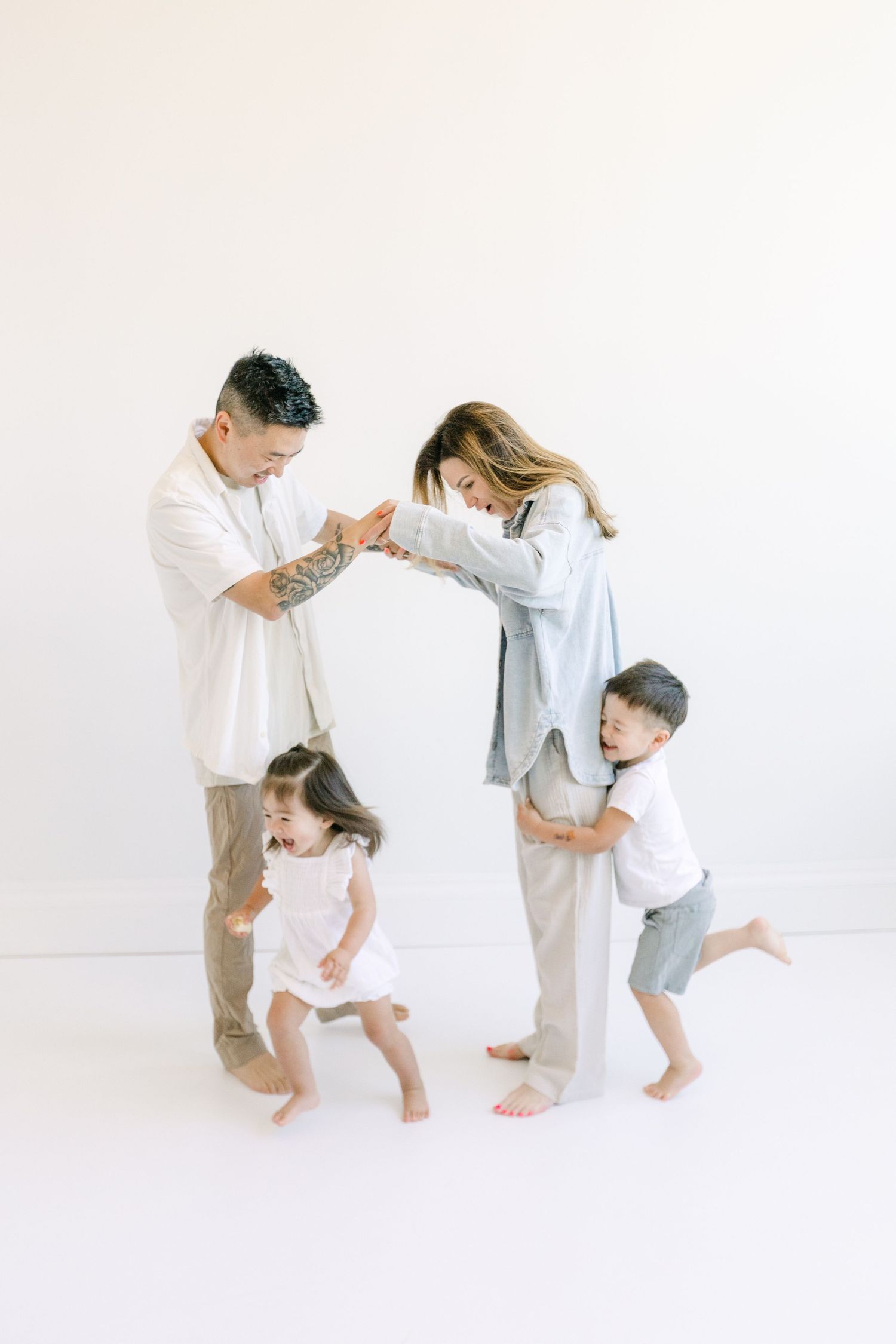 A joyful family of four plays together in a bright white studio, with parents and two young children dancing barefoot.