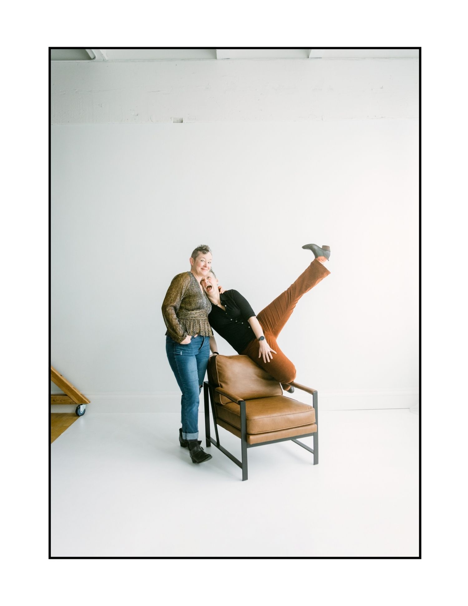 Two women laughing as one playfully flips upside down in a leather chair in a white photo studio.