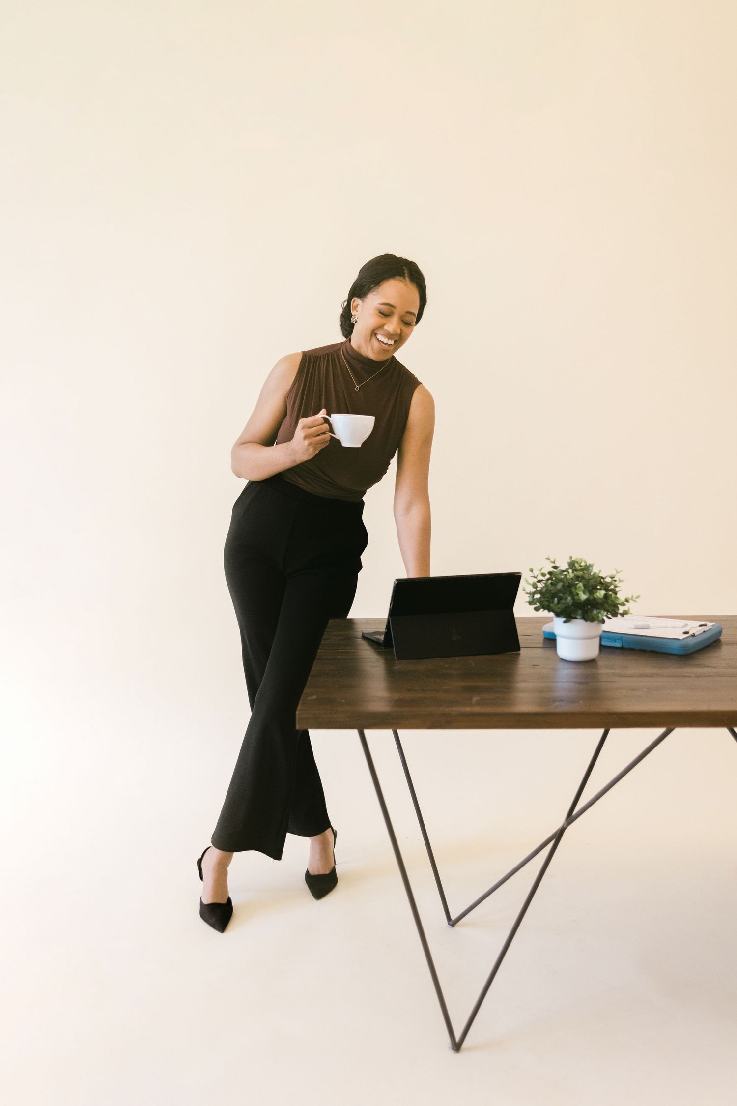 Professional woman in black outfit holding coffee cup, leaning against wooden desk with tablet and plant in bright office.