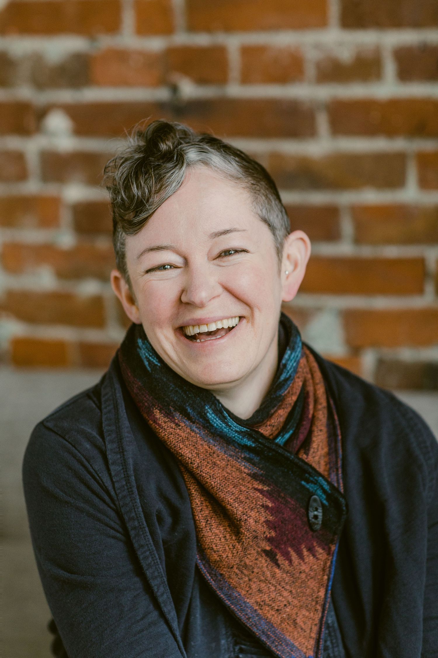 Smiling person with short hair wearing a colorful scarf and dark jacket, posing in front of a brick wall.