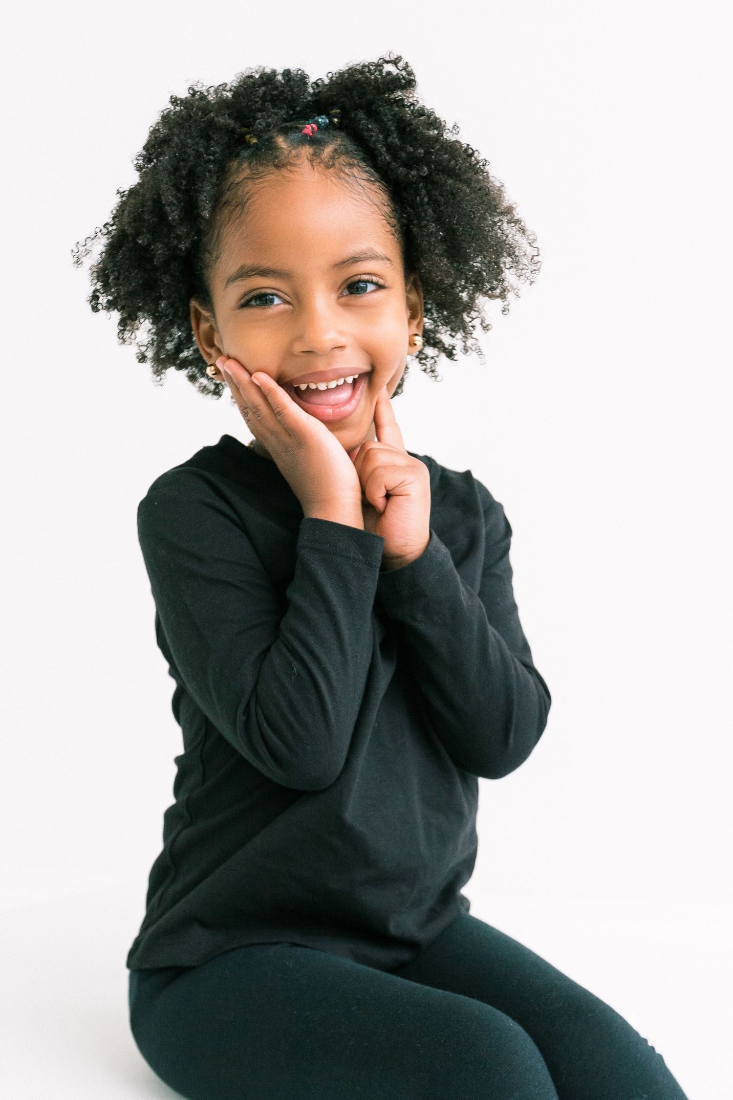 Adorable little girl in black outfit smiling with hands on cheeks against white background.