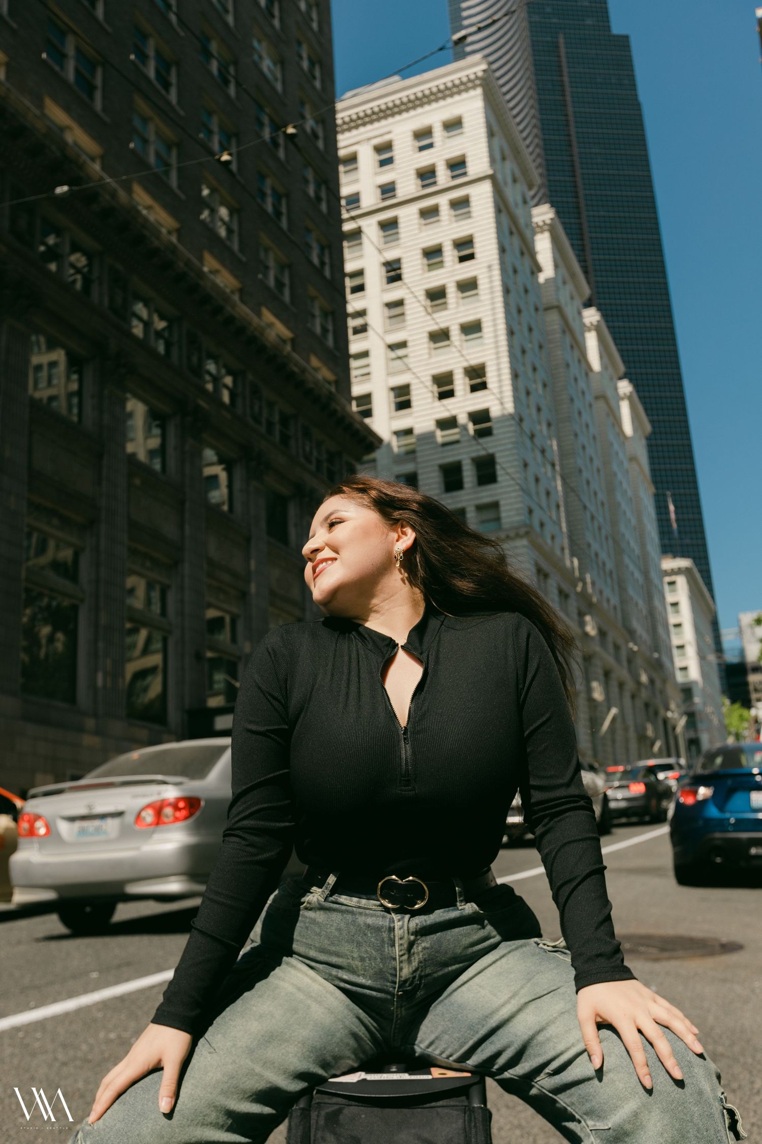 A smiling woman in a black zip-up top and jeans poses on a city street surrounded by tall skyscrapers.