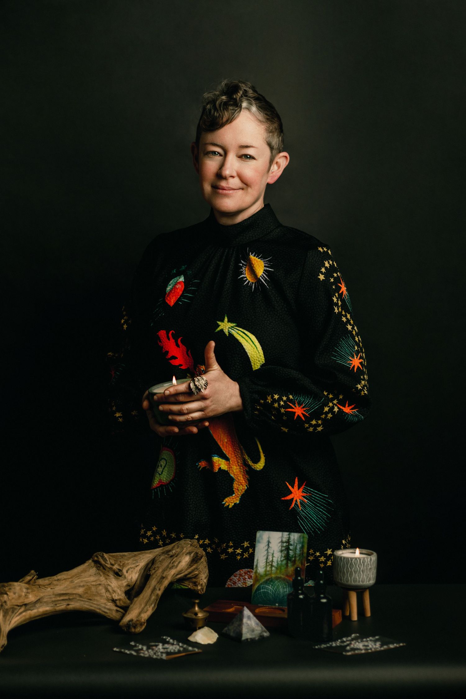 Woman in black celestial-print dress holding a candle, standing before crystals and driftwood in a dark studio.