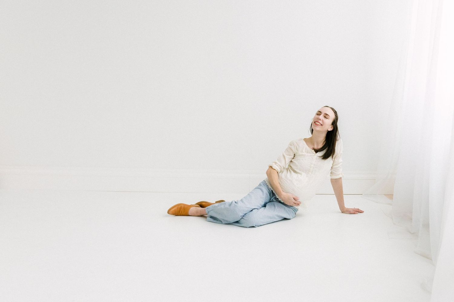 Pregnant woman sitting on white floor in jeans and cream top, smiling with head tilted back, cradling baby bump.