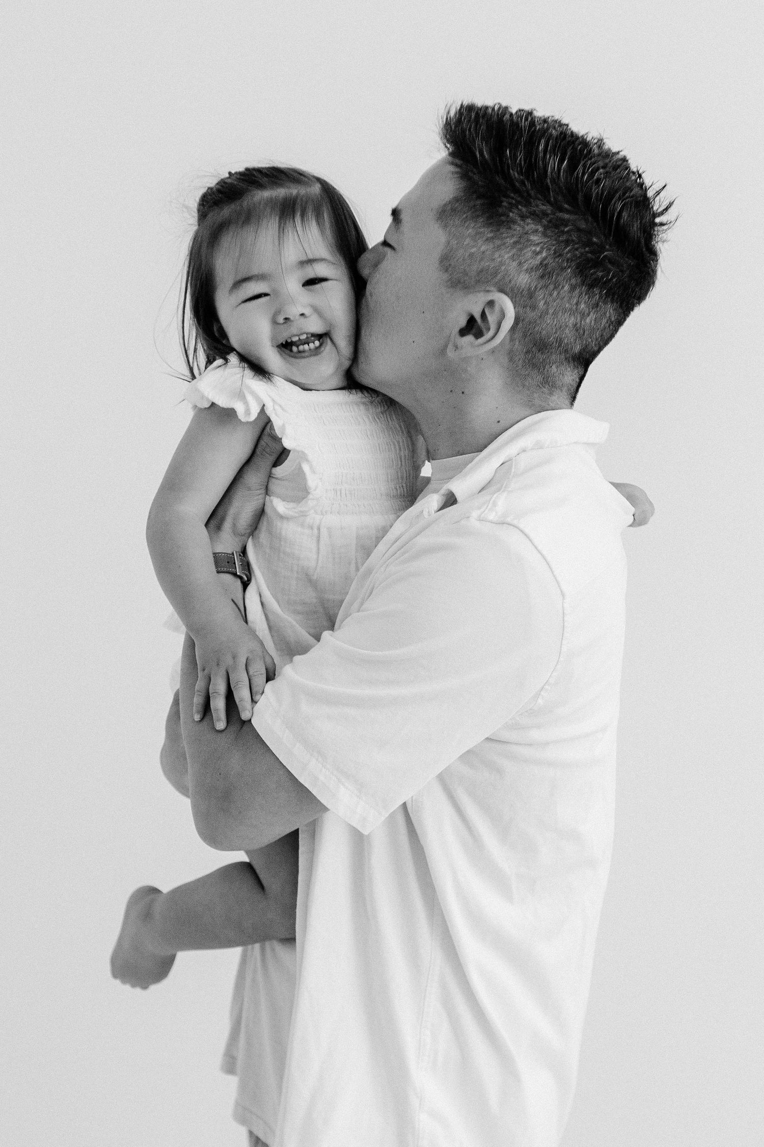 Black and white photo of a man kissing a laughing toddler girl he holds in his arms.