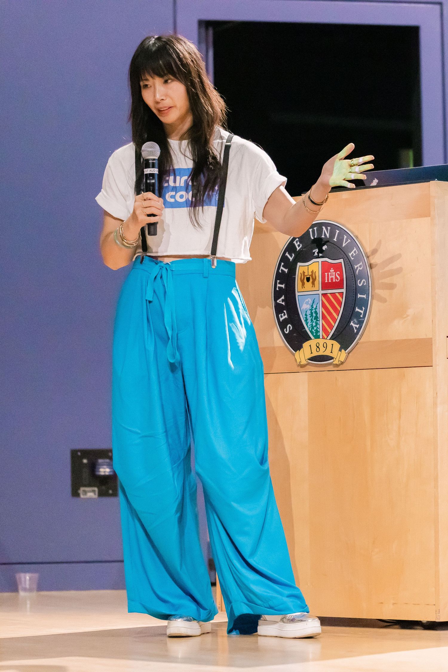 Young Asian woman in blue wide-leg pants speaking into microphone at Seattle University event.