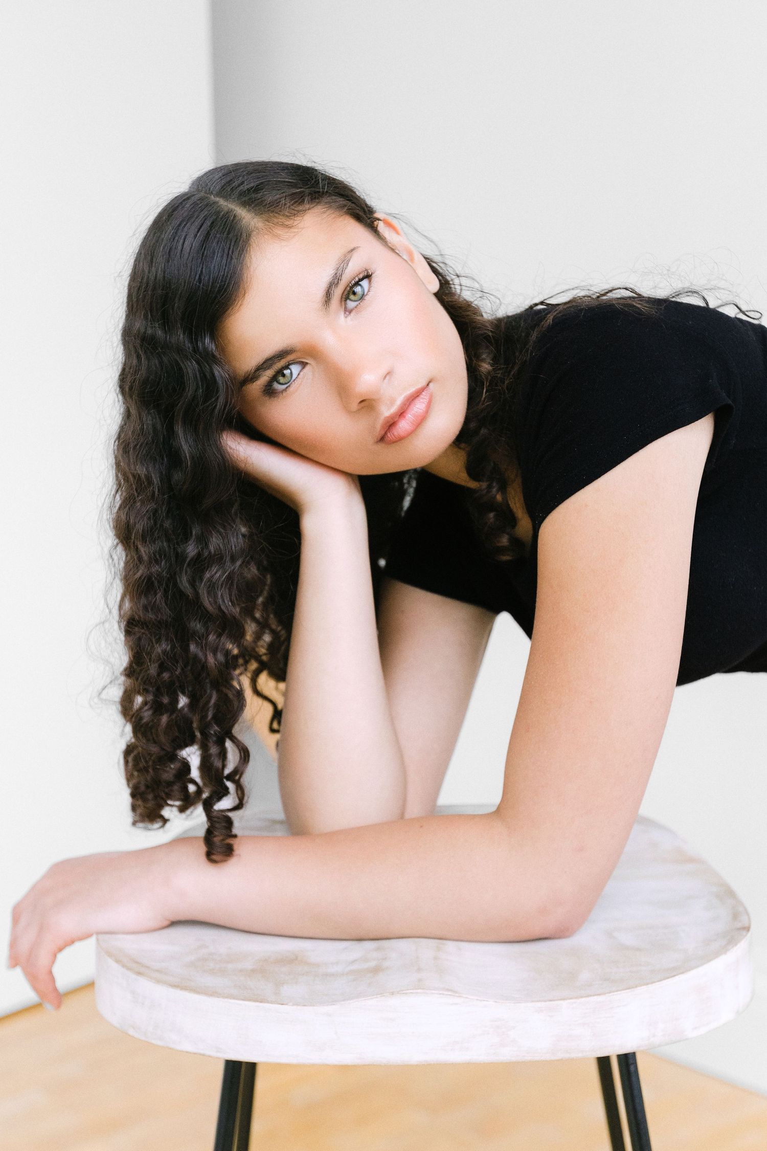 Young woman with curly dark hair resting her head on her hand while leaning on a white stool.