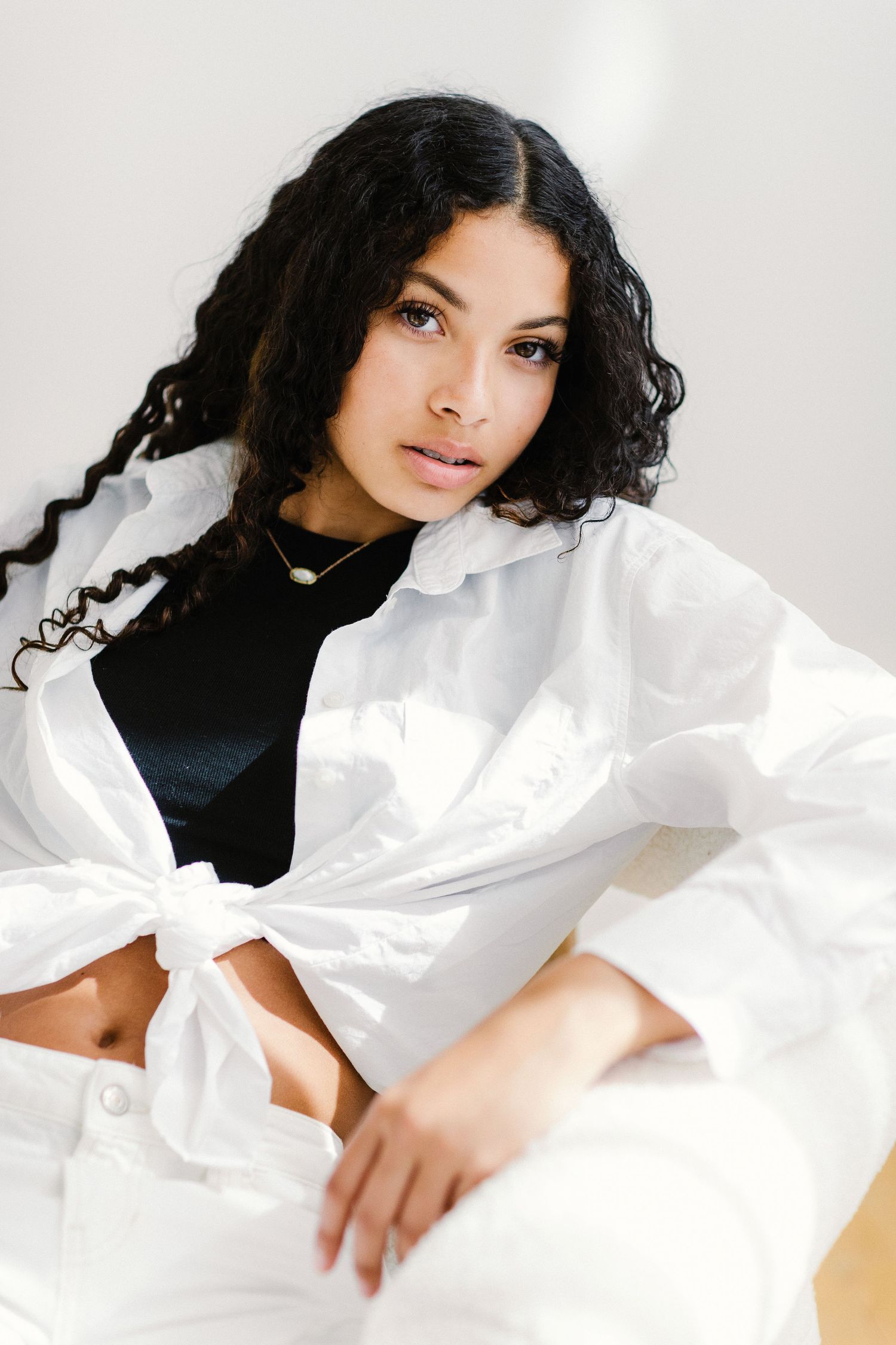 Young woman with curly black hair posing in white oversized shirt tied at waist over black crop top.