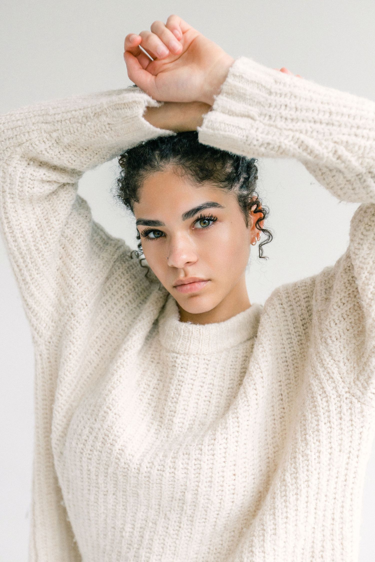 Young woman in cream knit sweater posing with arms raised, curly hair and natural makeup against white background.
