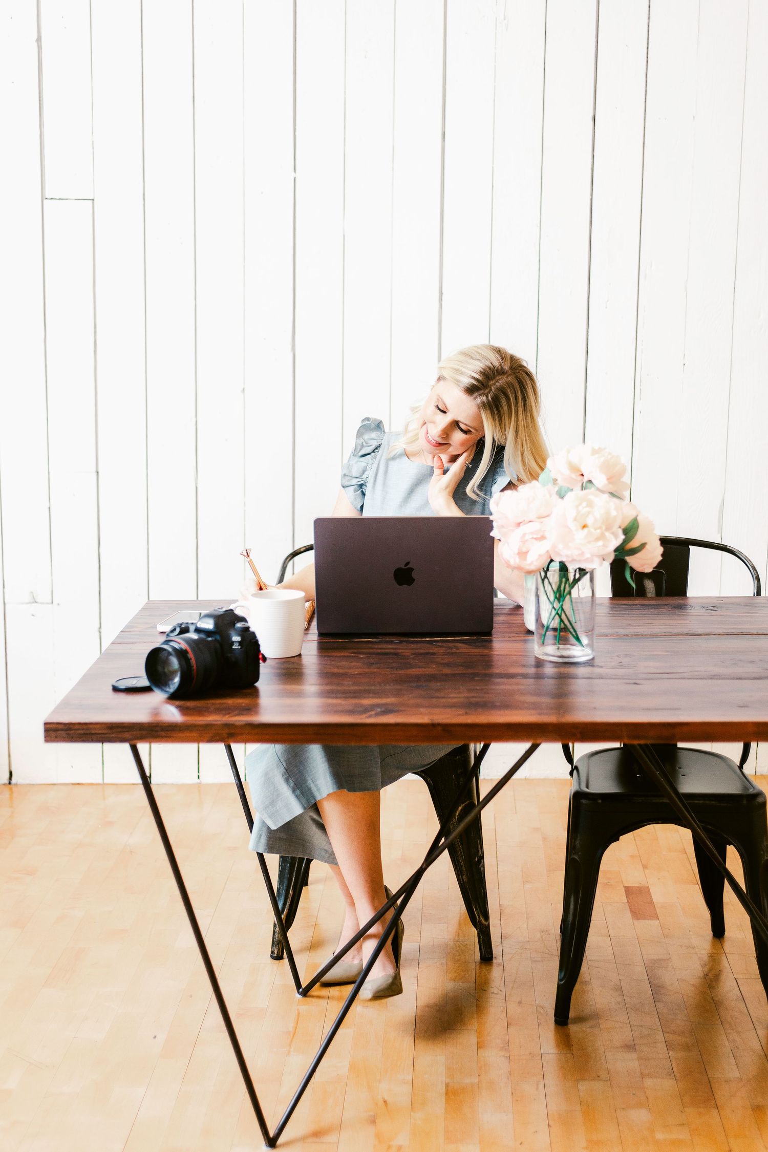 Blonde woman smiling at MacBook laptop on wooden desk with camera and pink flowers in bright white office.