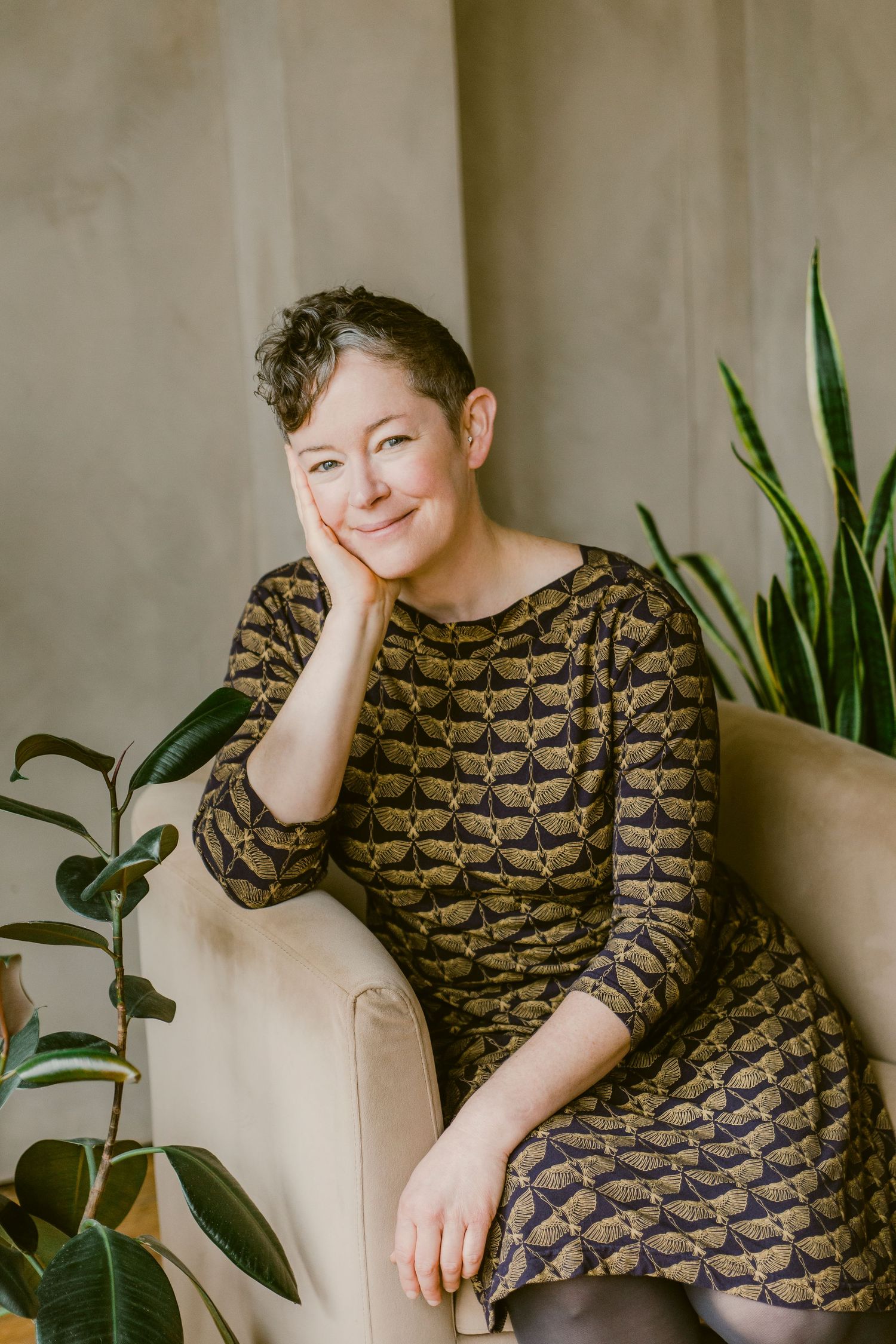 Smiling woman with short hair in patterned dress seated on beige chair surrounded by green indoor plants.