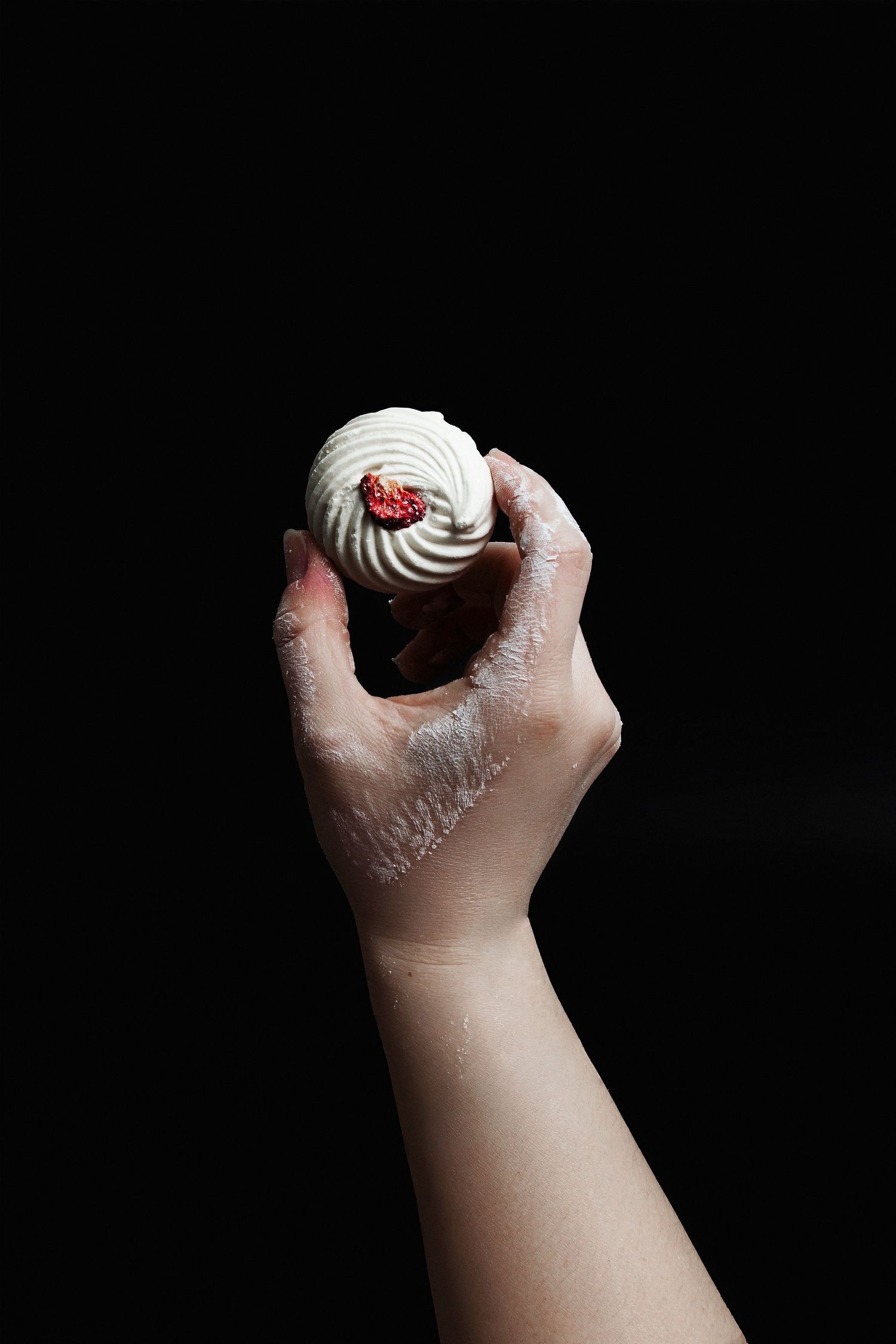 Hand holding a white cream dessert ball topped with a red raspberry against a black background.