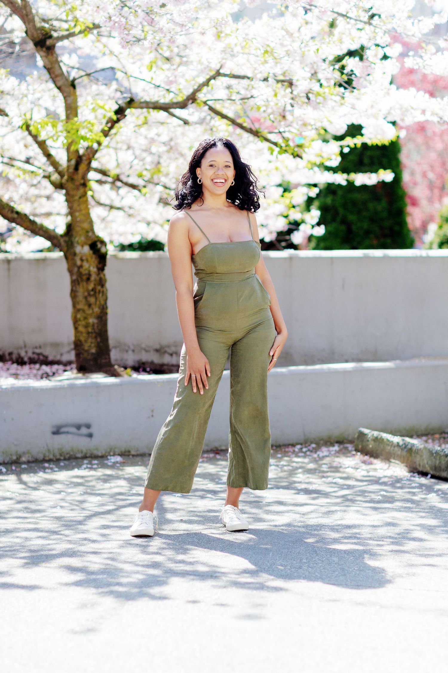 Young woman in olive green jumpsuit smiling outdoors near cherry blossom trees in spring.