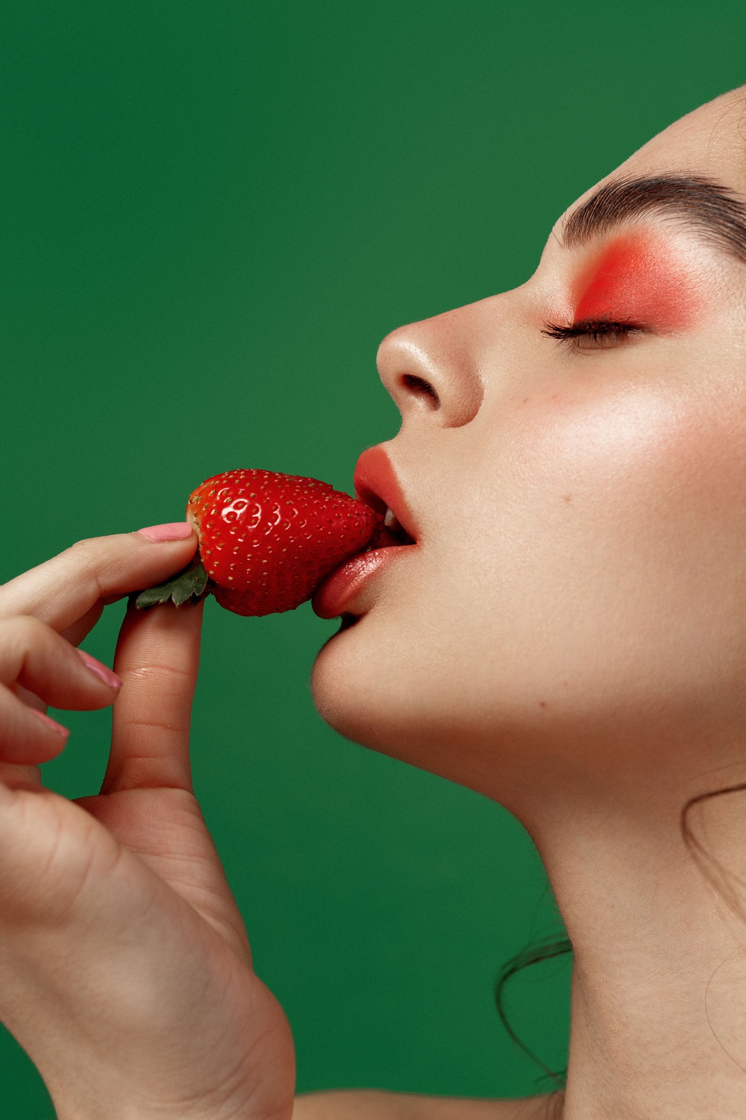 Close-up beauty shot of woman with red eyeshadow and lips biting into a fresh red strawberry on green background.
