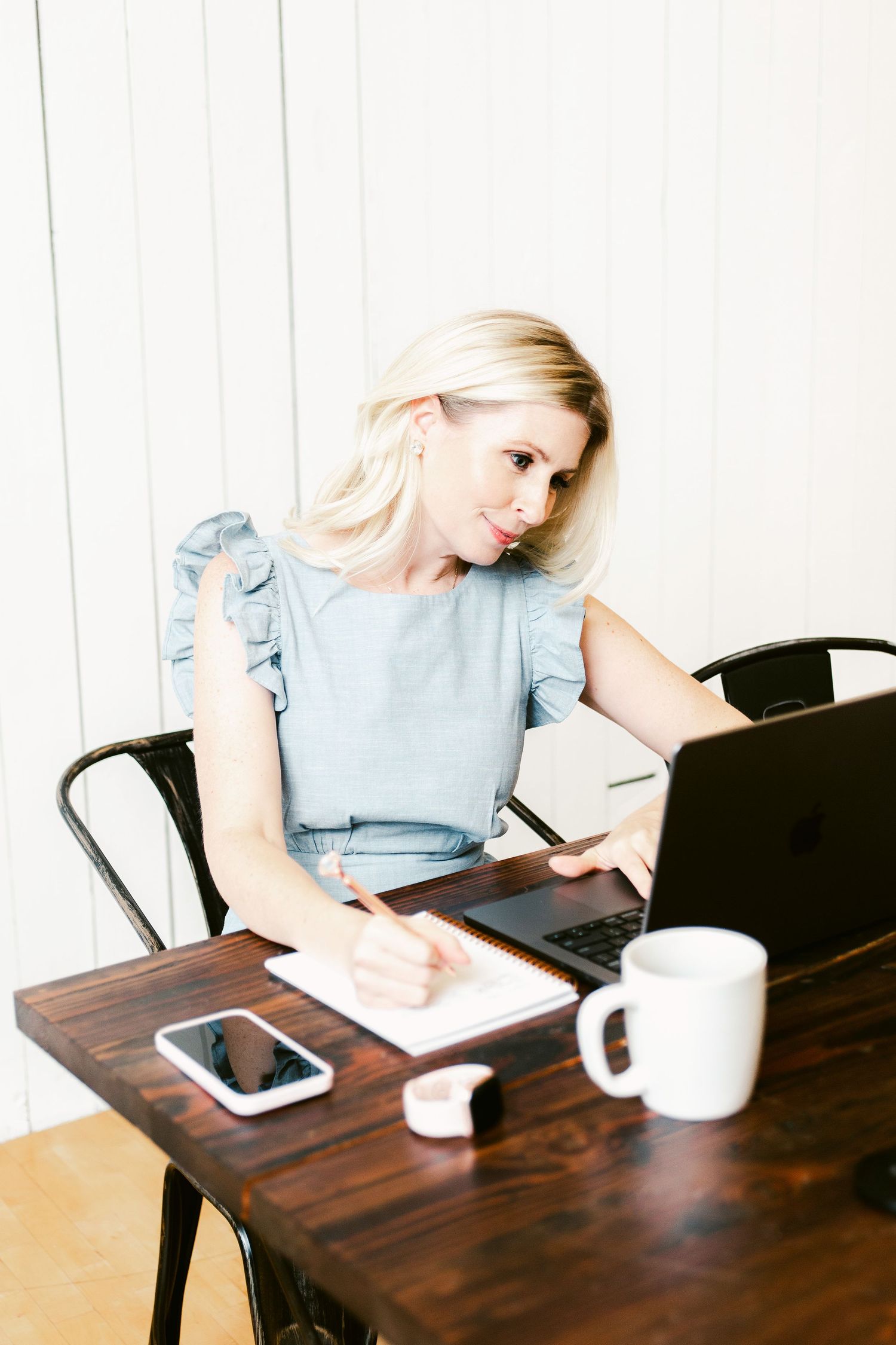 Blonde woman in blue ruffled top writing in notebook at wooden desk with laptop, phone, and coffee mug.
