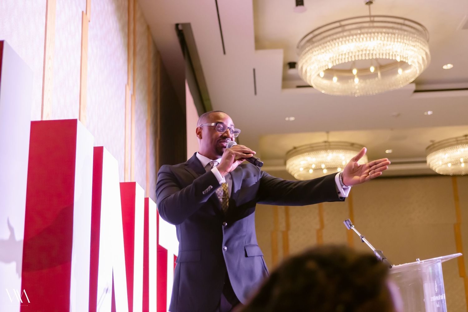 Black man in suit speaking passionately at podium during formal indoor conference event.