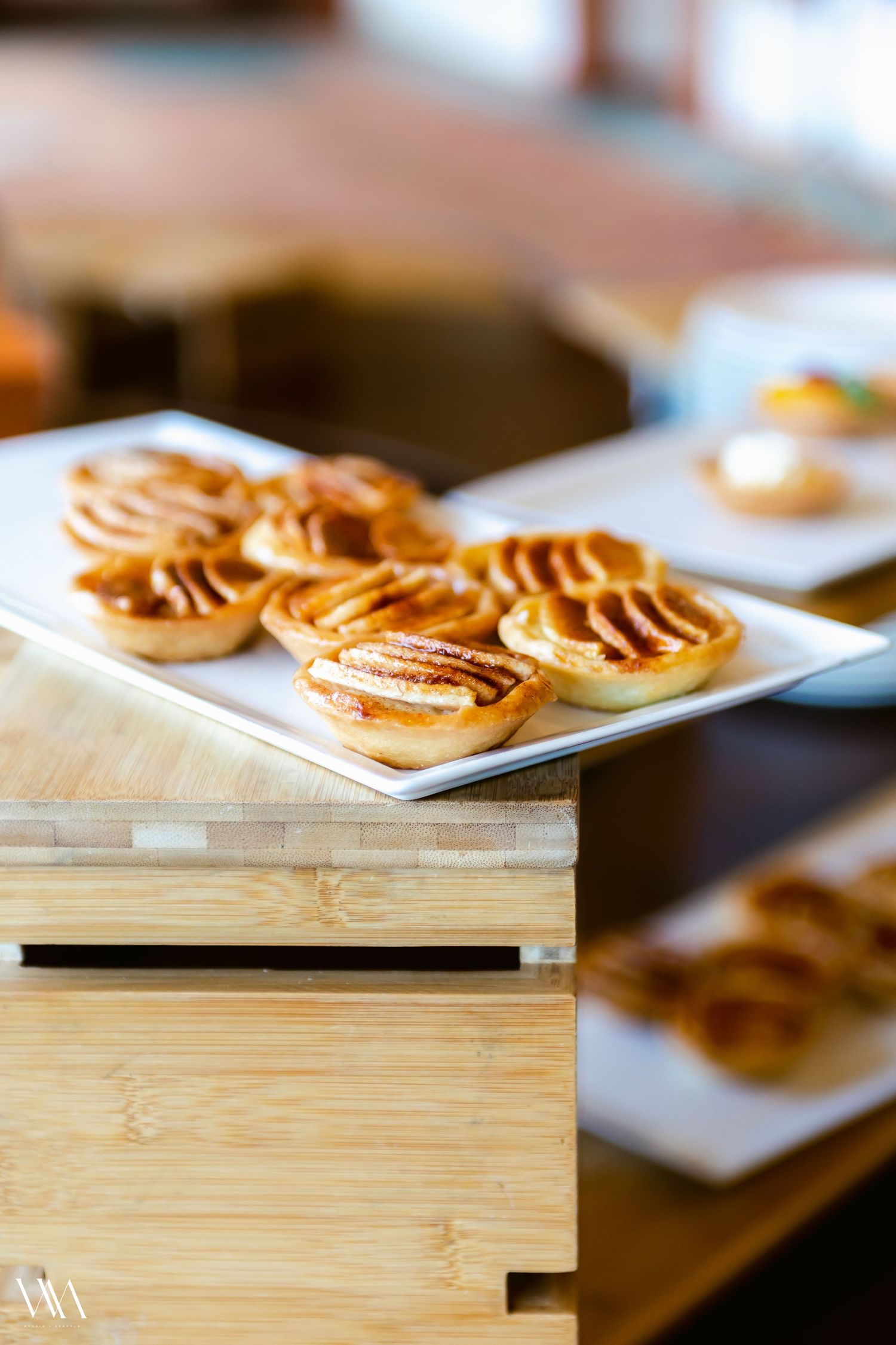 Golden apple tarts on a white plate atop a wooden display box at a bakery or catered event.