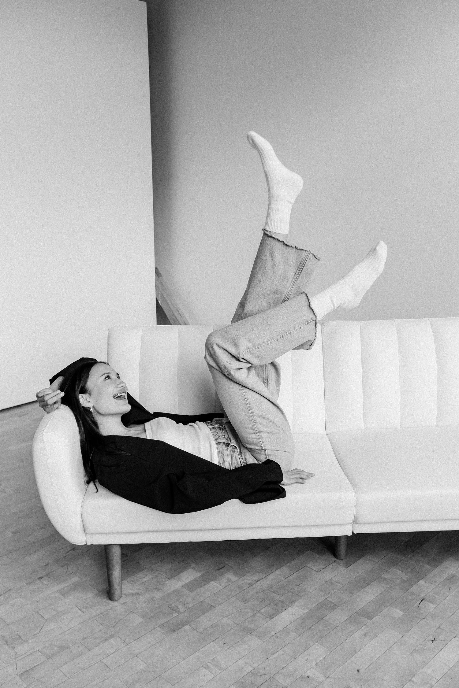 A black and white photo of a laughing woman lying on a white sofa with her legs raised in the air.