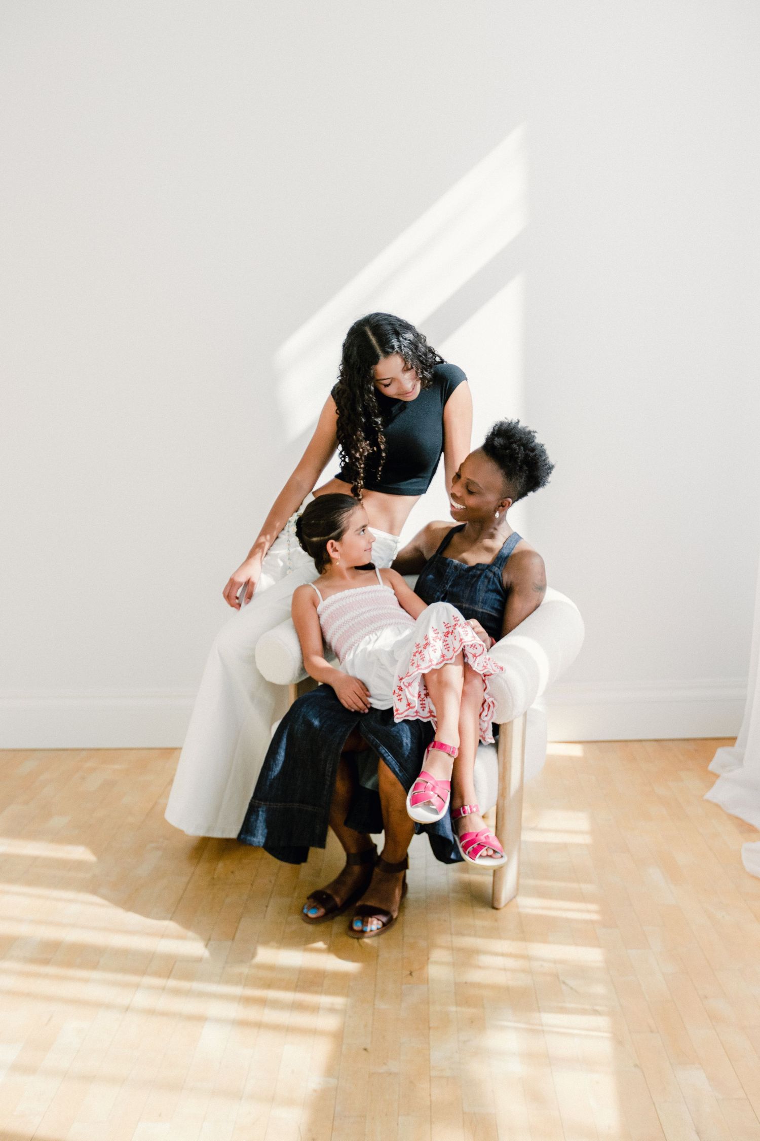 Two women and a young girl laugh together while seated in a white chair in a sunlit studio.