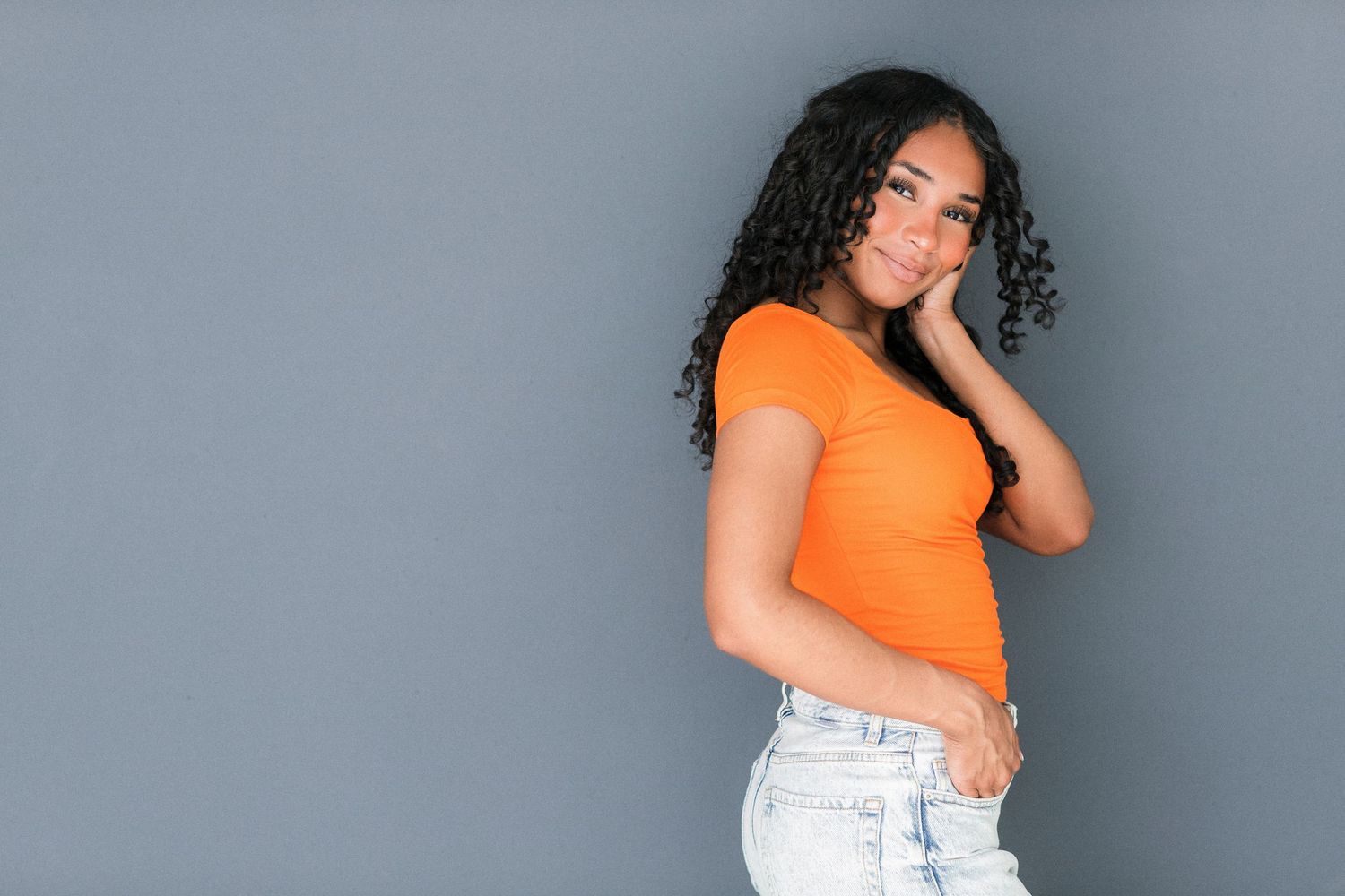 Young woman in orange top and jeans posing against gray background, smiling with hand on cheek.