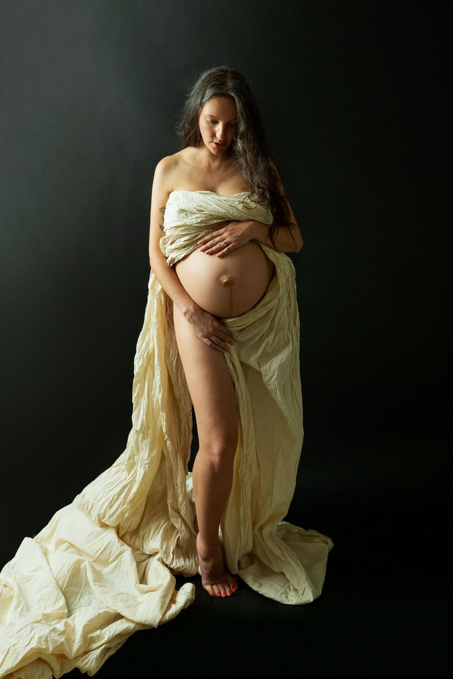 Pregnant woman draped in flowing cream fabric poses gracefully against dark background, cradling her bare baby bump.