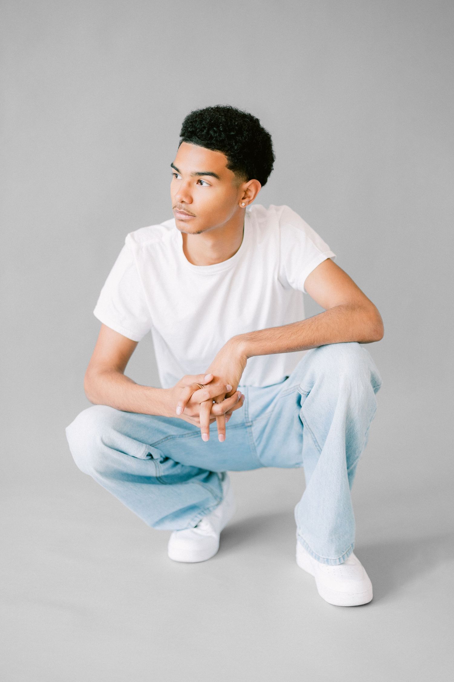 Young Black man in white t-shirt and light blue jeans crouching on gray background, looking sideways.