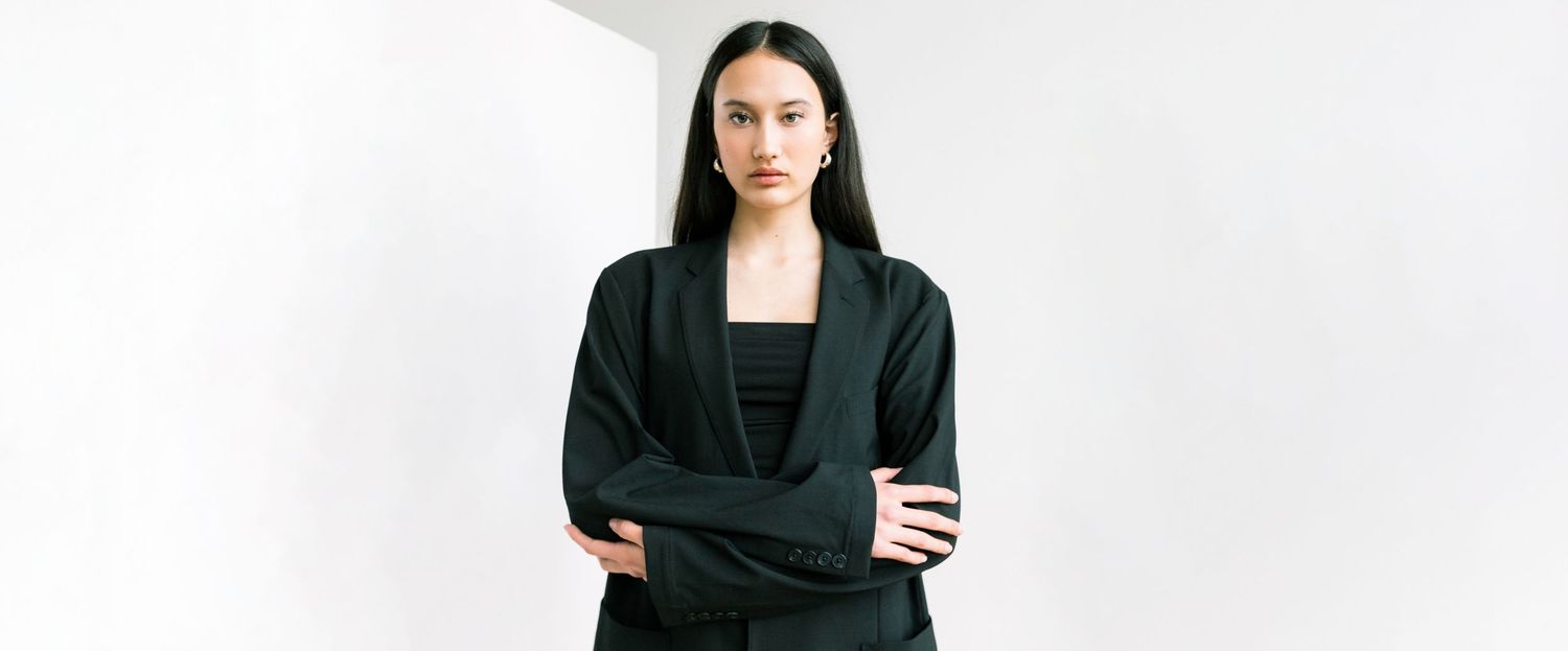 Woman in black blazer and top stands with arms crossed against white background, exuding confidence.