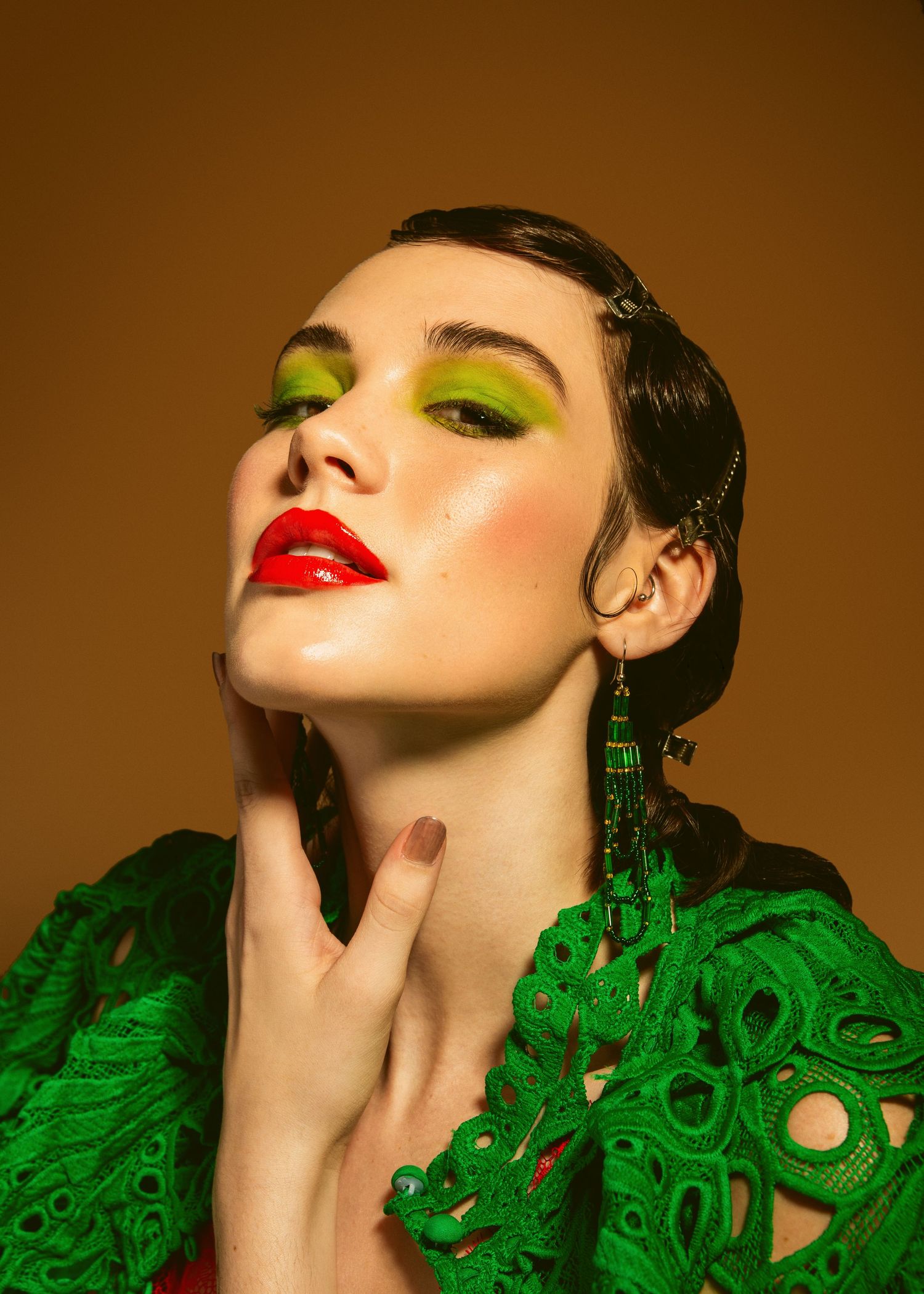 Woman with bold green eyeshadow, red lips, long beaded earrings, wearing a green textured outfit against a warm background.