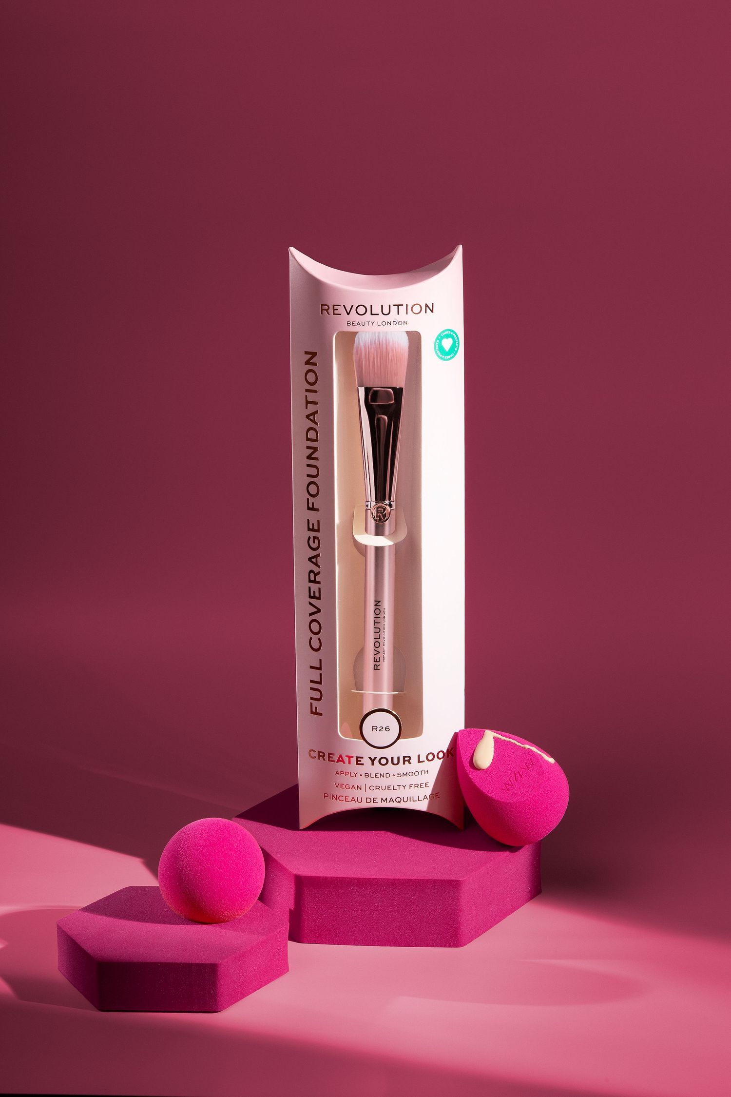 Revolution Full Coverage Foundation brush in pink packaging displayed with hot pink beauty sponges on a pink background.