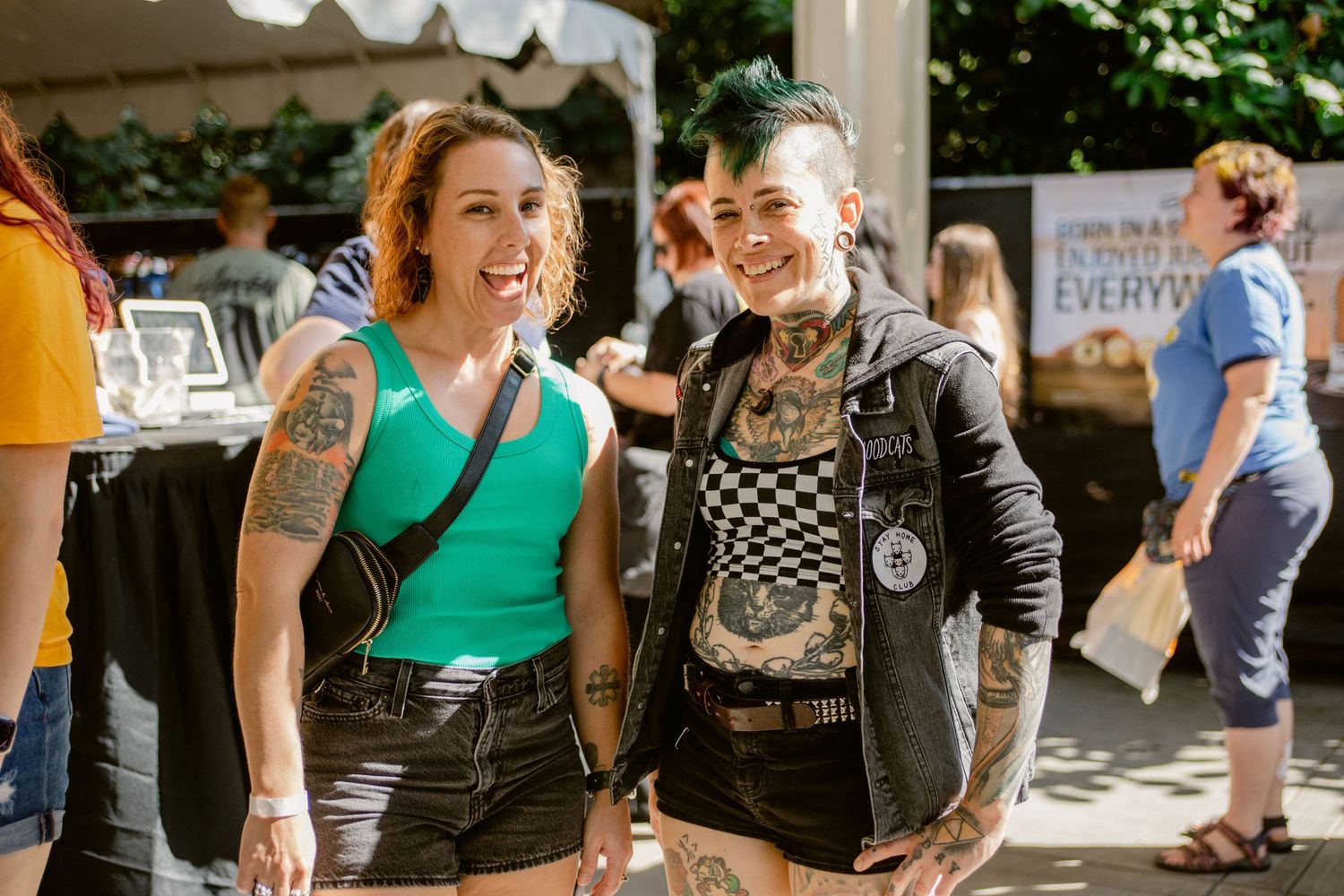 Two tattooed women smiling together at outdoor festival marketplace event.